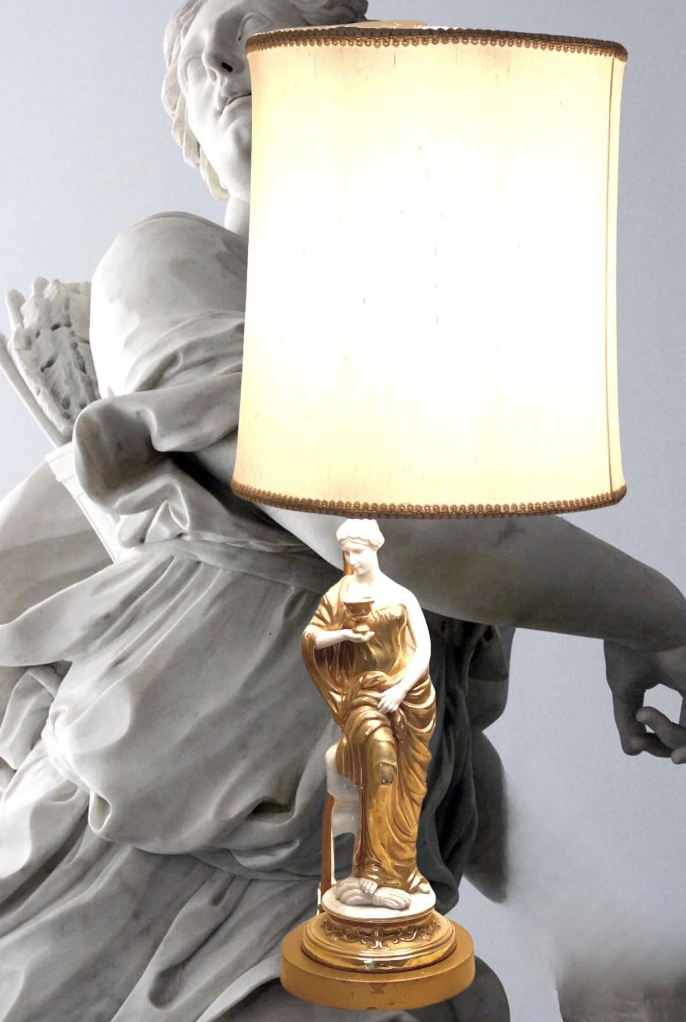 A decorative table lamp with a classical statue base and a large lampshade, placed in front of a marble statue of a seated figure holding an object, blending classical art and modern decor.