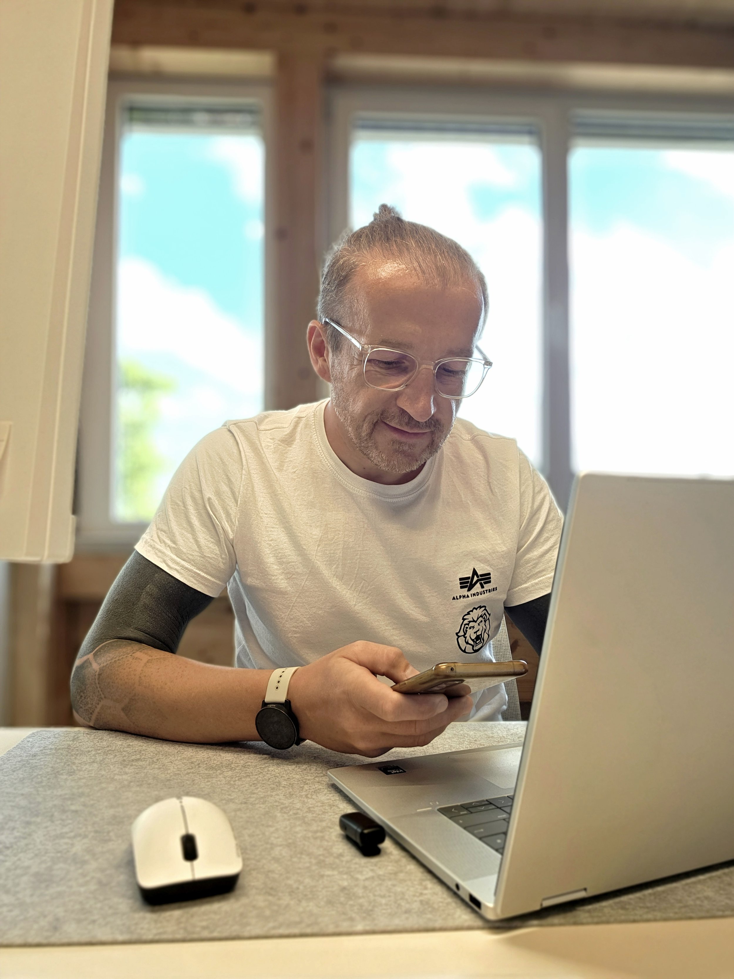 Mann mit Brille, im weißen T-Shirt mit Logos, sitzt an einem Tisch, schaut auf sein Smartphone, im Hintergrund große Fenster mit blauer Himmel und Wolken, Arbeitsplatz mit Laptop, Maus und kleinen Gegenständen.