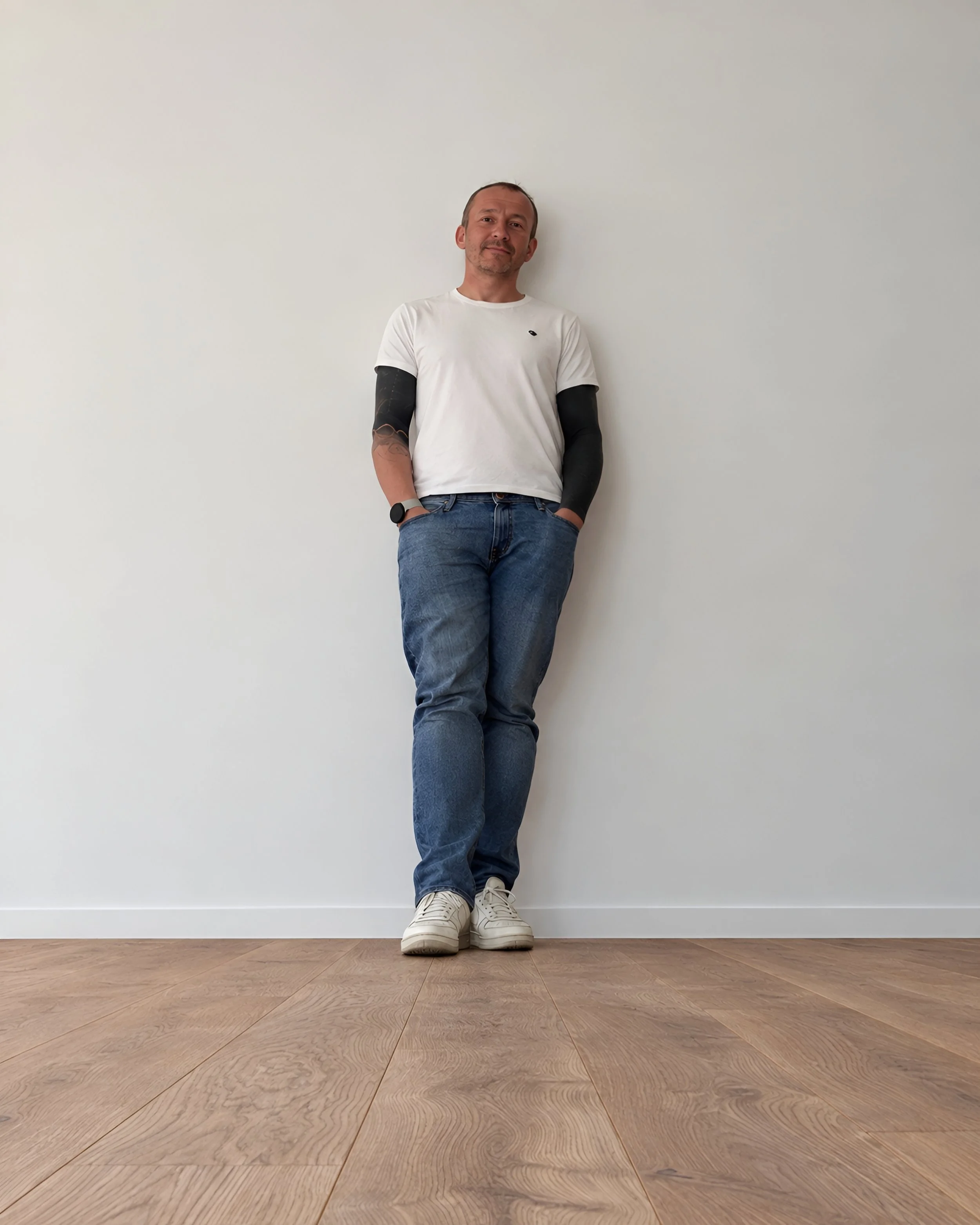 Mann in weißem T-Shirt, Jeans und weißen Sneakers, steht vor einer weißen Wand im Raum.