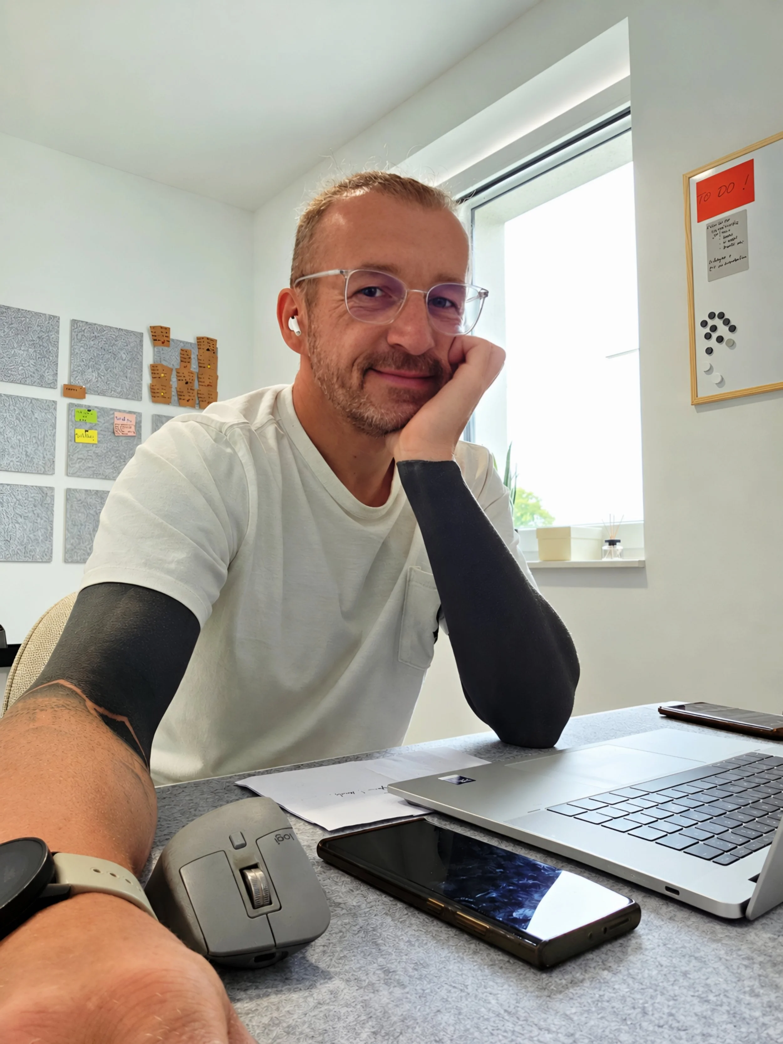 Ein Mann sitzt an einem Schreibtisch mit einem Laptop, einem Smartphone, einem Tablet. Er trägt eine Brille, AirPods und hat einen Bart. Im Hintergrund eine weiße Wand mit Notizzetteln und Postern.