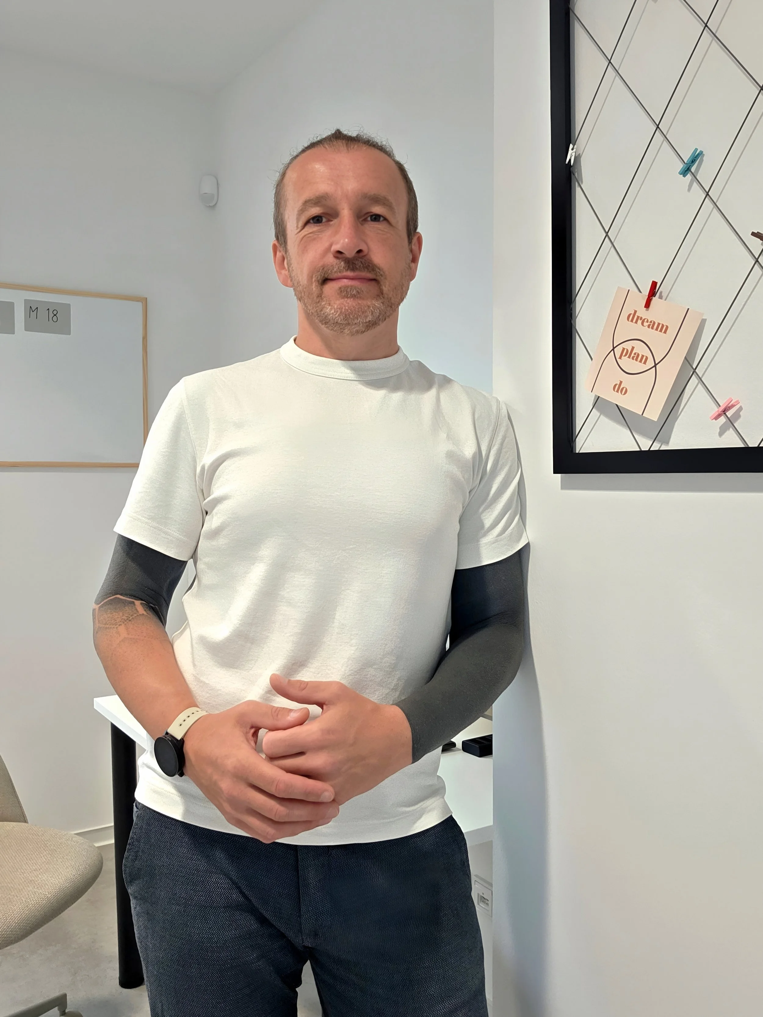 Ein Mann mit kurzen Haaren und Bart steht in einem Raum, trägt ein weißes T-Shirt und dunkle Hosen, trägt eine Armbanduhr und hat Tattoos am rechten Arm.