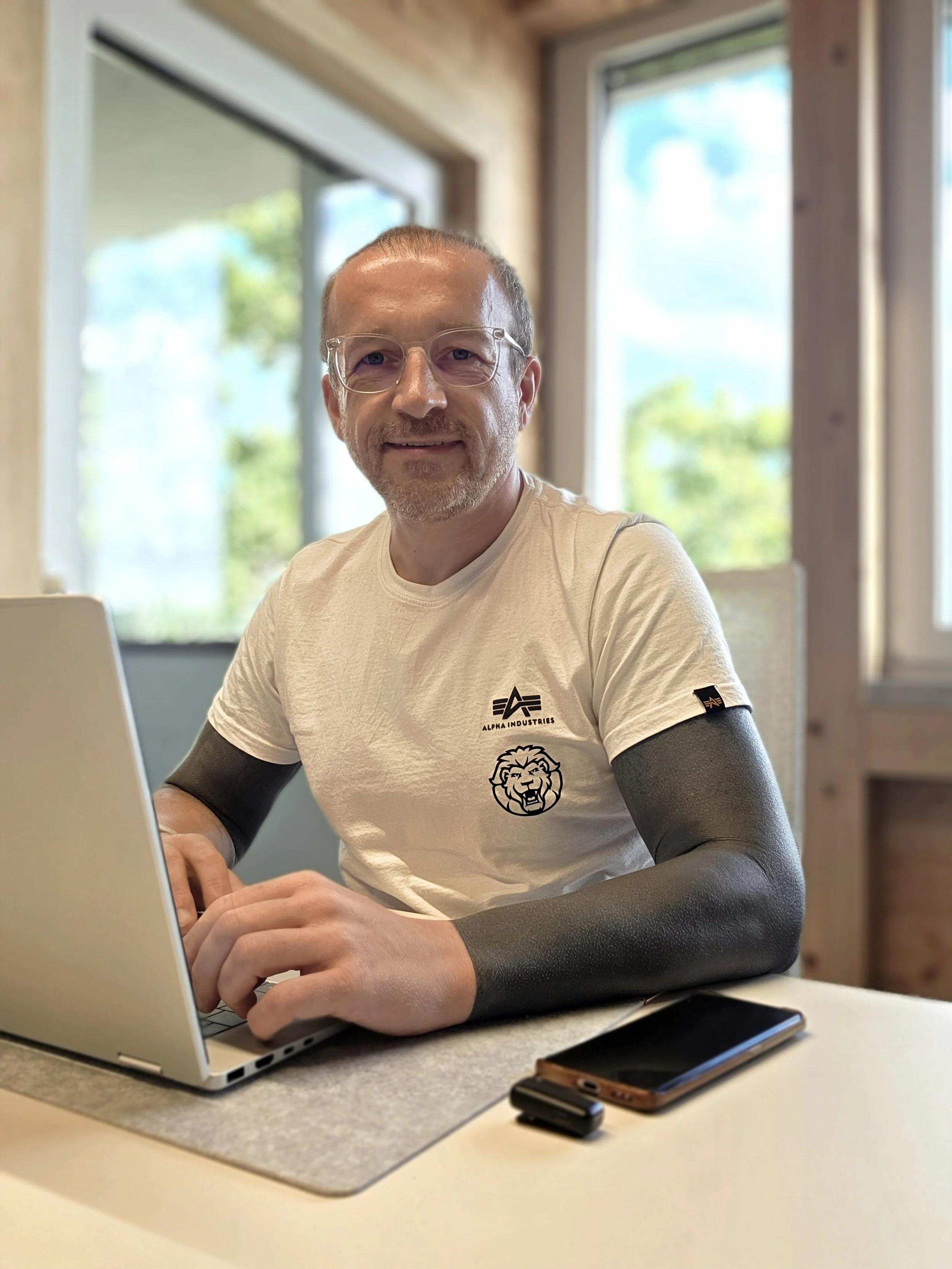 Mann mit Brille und Bart sitzt an einem Tisch mit Laptop, Smartphone, im Hintergrund Fenster mit Blick ins Grüne, innen mit Holz verziert.