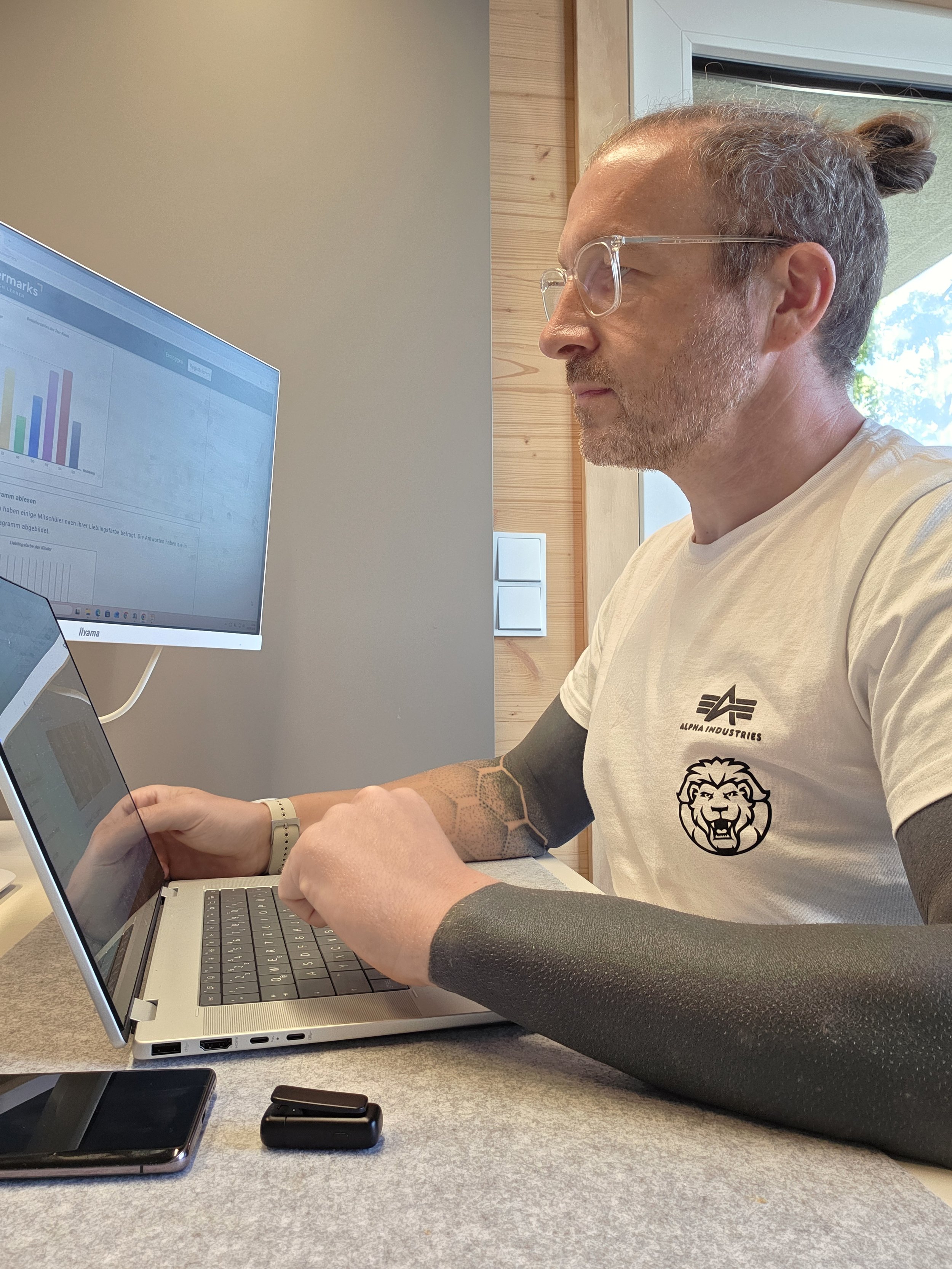 Ein Mann arbeitet an einem Laptop in einem Büro. Er trägt ein weißes T-Shirt mit einem Löwen-Logo und der Text 'ALPHA INDUSTRIES'.