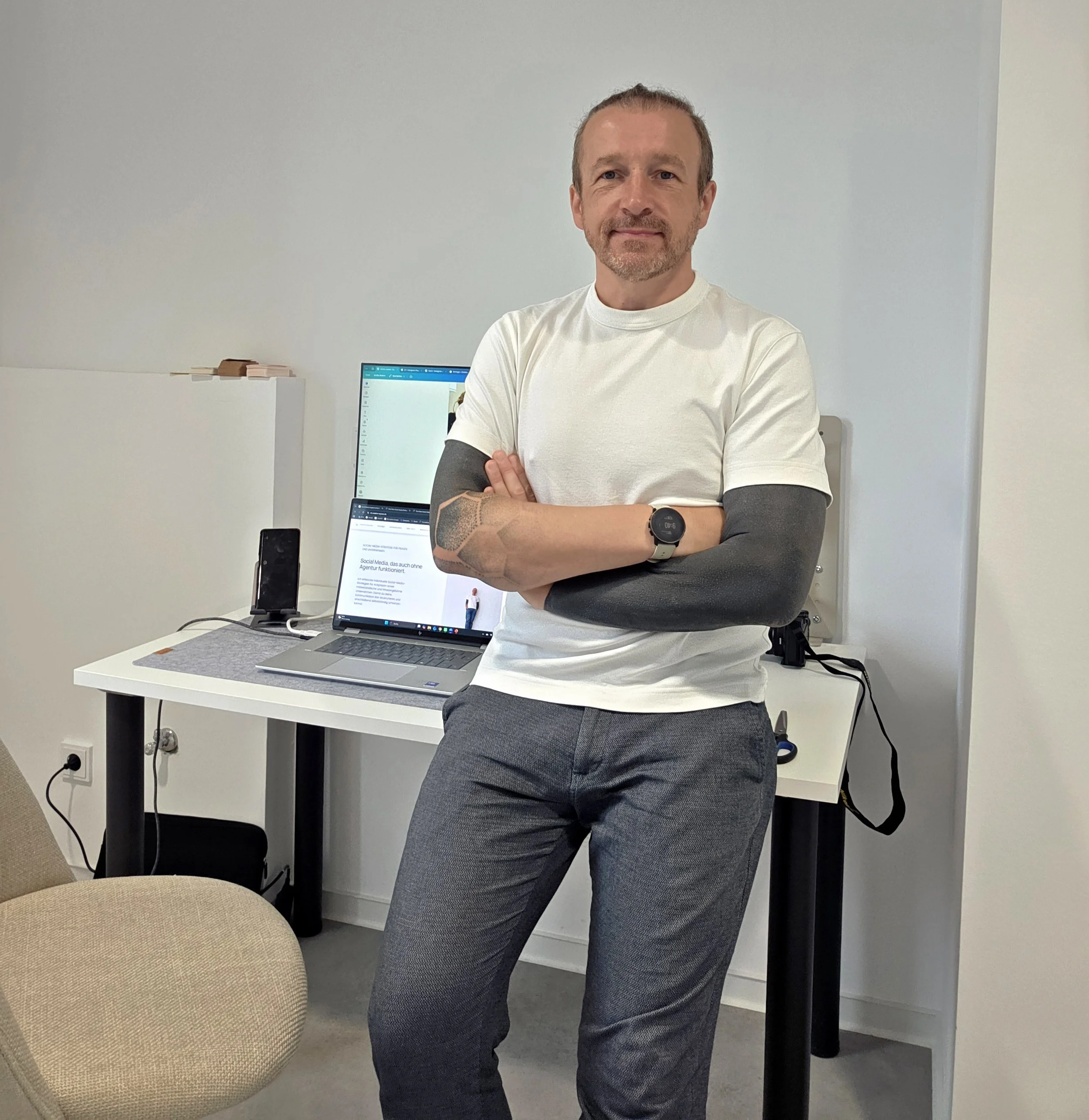 Ein Mann mit kurzen, braunen Haaren und Bart trägt ein weißes T-Shirt in einem modernen Büro. Er steht mit verschränkten Armen vor einem Bildschirm, der eine Webseite anzeigt, und auf einem Tisch neben ihm liegen ein Smartphone und ein Notizbuch.