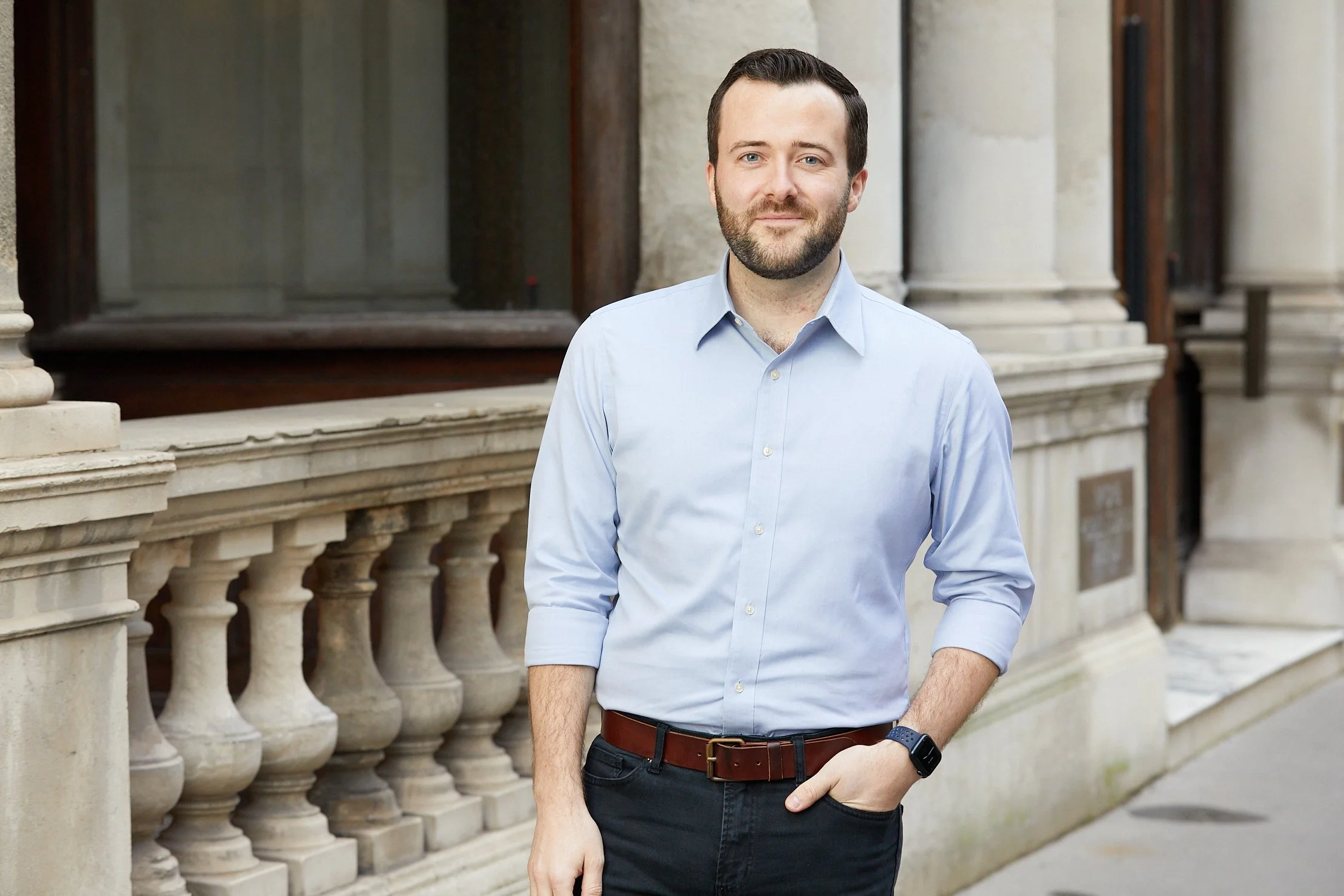 A man with dark hair and a beard standing outdoors in front of a stone building with classical columns, wearing a light blue button-up shirt, dark pants, a belt, and a smartwatch, with one hand in his pocket.