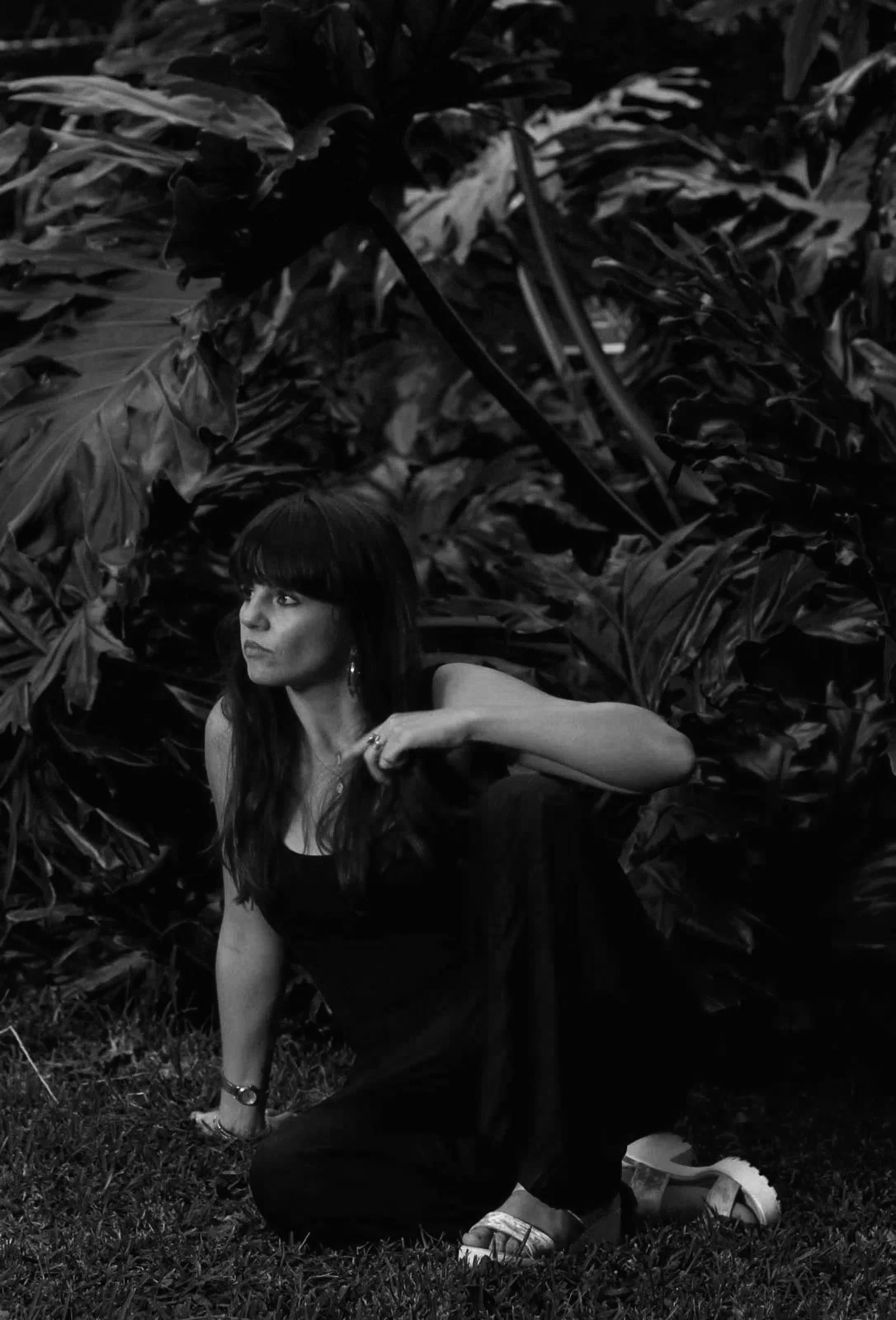 A woman with long dark hair and bangs, wearing a black sleeveless top and black pants, is crouching outdoors on grass, surrounded by large tropical leaves, with her left arm resting on her knee and her right hand near her shoulder, looking to her left.