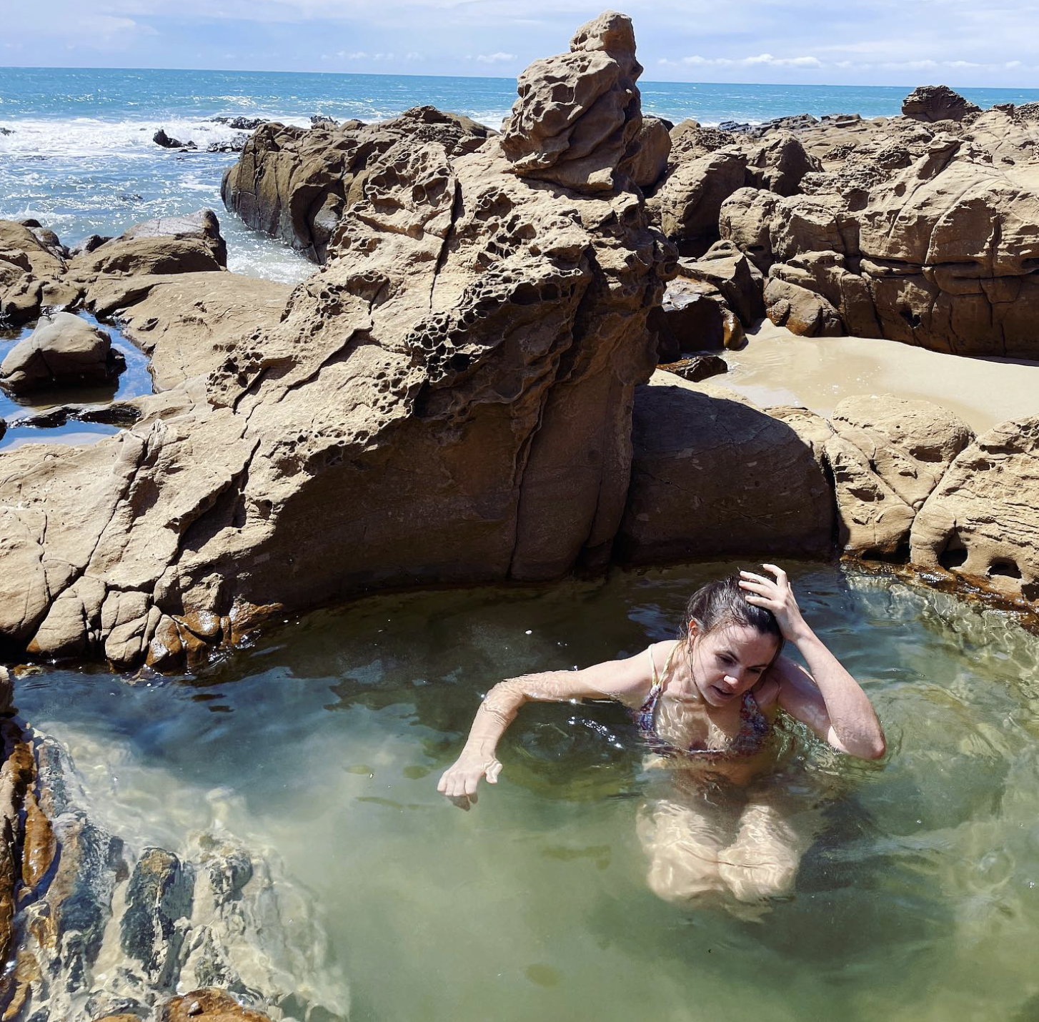 A woman swimming in a rocky ocean pool with large rocks and the sea in the background.
