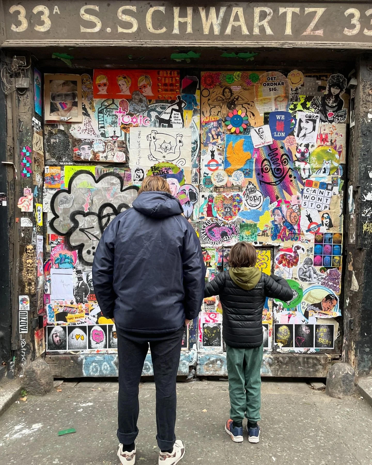 Two people, an adult and a child, look at a wall covered in colorful posters and stickers. The adult is wearing a dark jacket and the child is wearing a black jacket and green pants. They are standing in front of a graffiti-filled entrance labeled with the address 33 S. Schwartz.