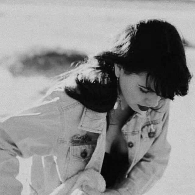 Black and white photo of a woman with dark hair, dressed in a denim jacket, looking down, possibly at the beach or outdoors.
