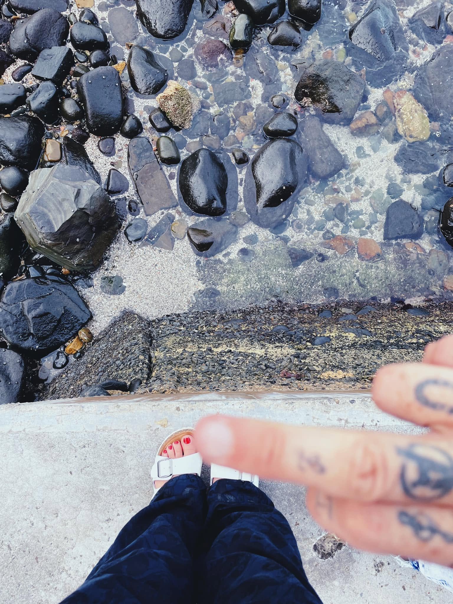 View of a person taking a photo from a high perspective, standing on a concrete surface near a rocky shoreline with water below, wearing flip-flops and dark clothing.