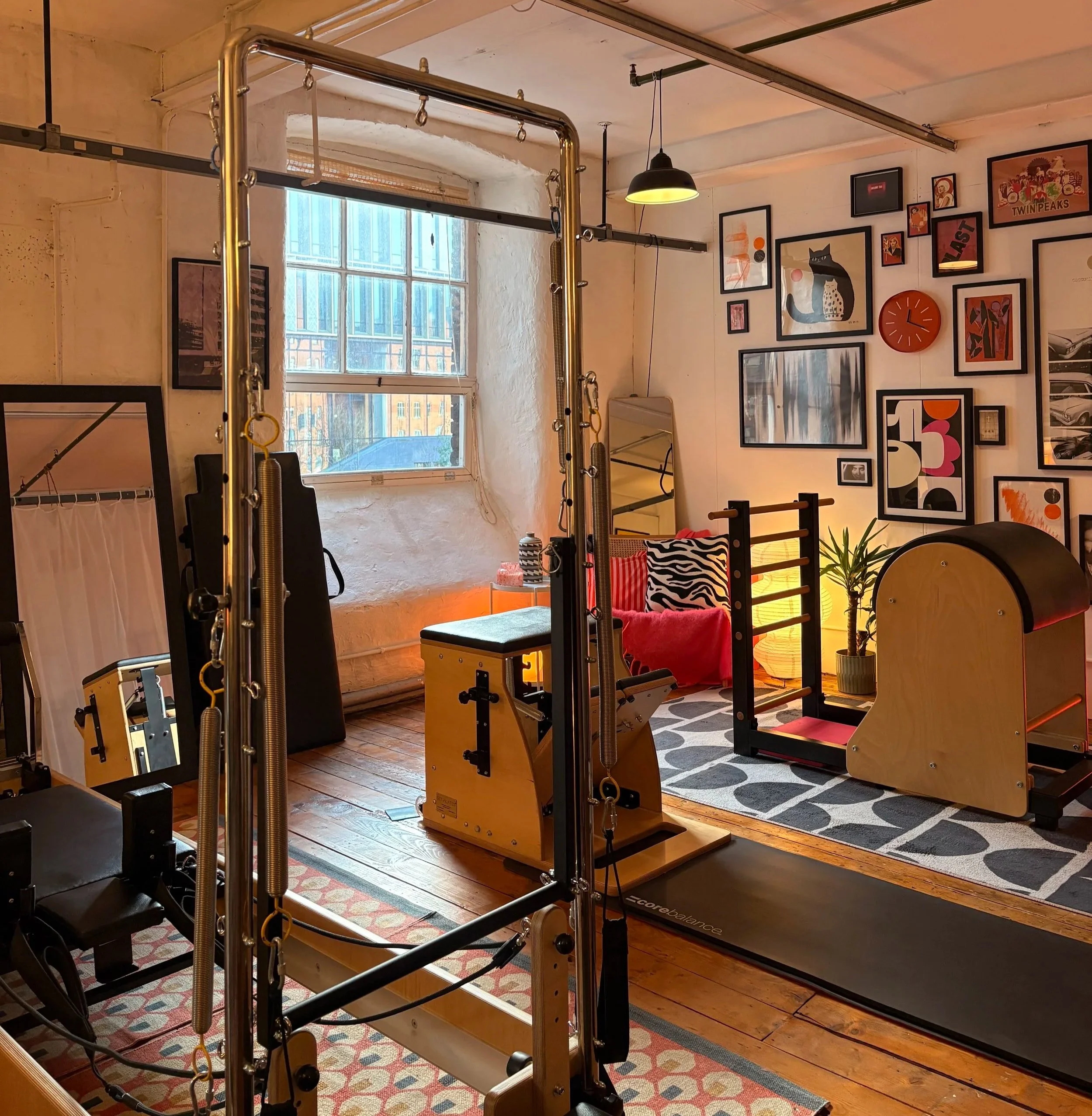 Indoor Pilates studio with various equipment, framed artwork on the wall, window letting in natural light, and a patterned rug on wooden floor.