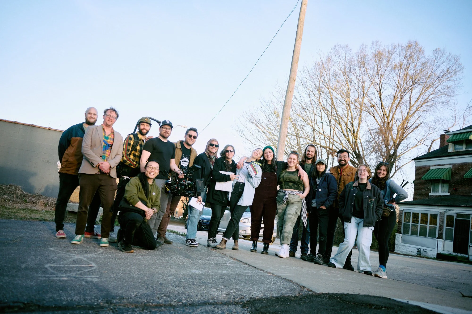 Group of diverse people standing outdoors on a sidewalk, some members are smiling and posing for the photo, with a large tree and a house in the background, under a clear sky.
