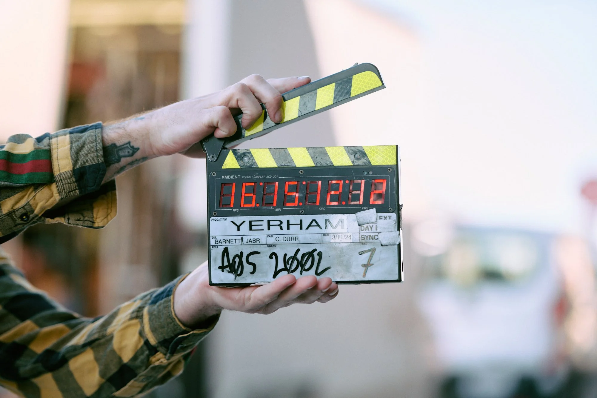 A person with a tattoo on their arm holding a clapperboard during filming, with the slate displaying the production name "YERHAM" and handwritten scene details, set outdoors with a blurred background.