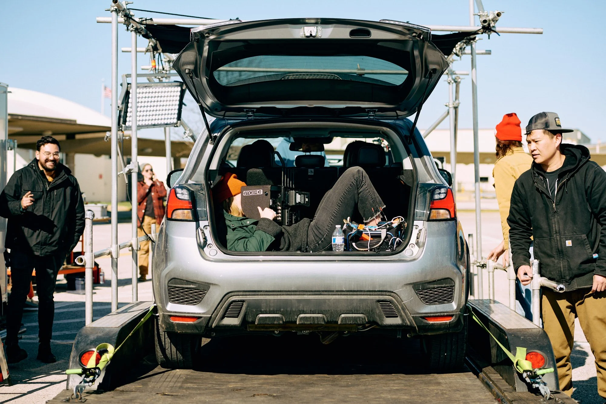 A person in a green jacket and black pants is lying inside a car's open trunk, holding a camera or filming equipment. Several other people are around, some smiling and taking photos, on a setup with scaffolding and equipment outdoors on a clear day.