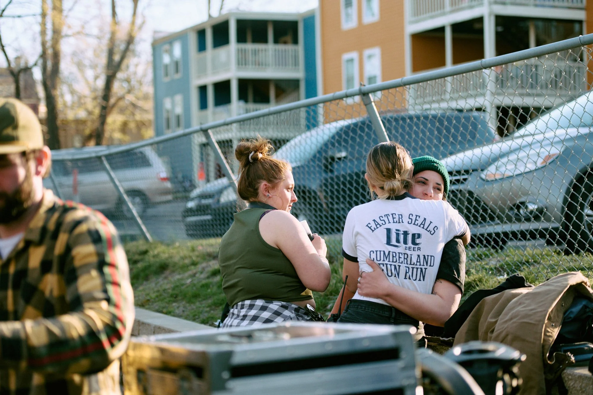 People sitting on the ground behind a chain-link fence in a park, with multistory apartment buildings in the background.