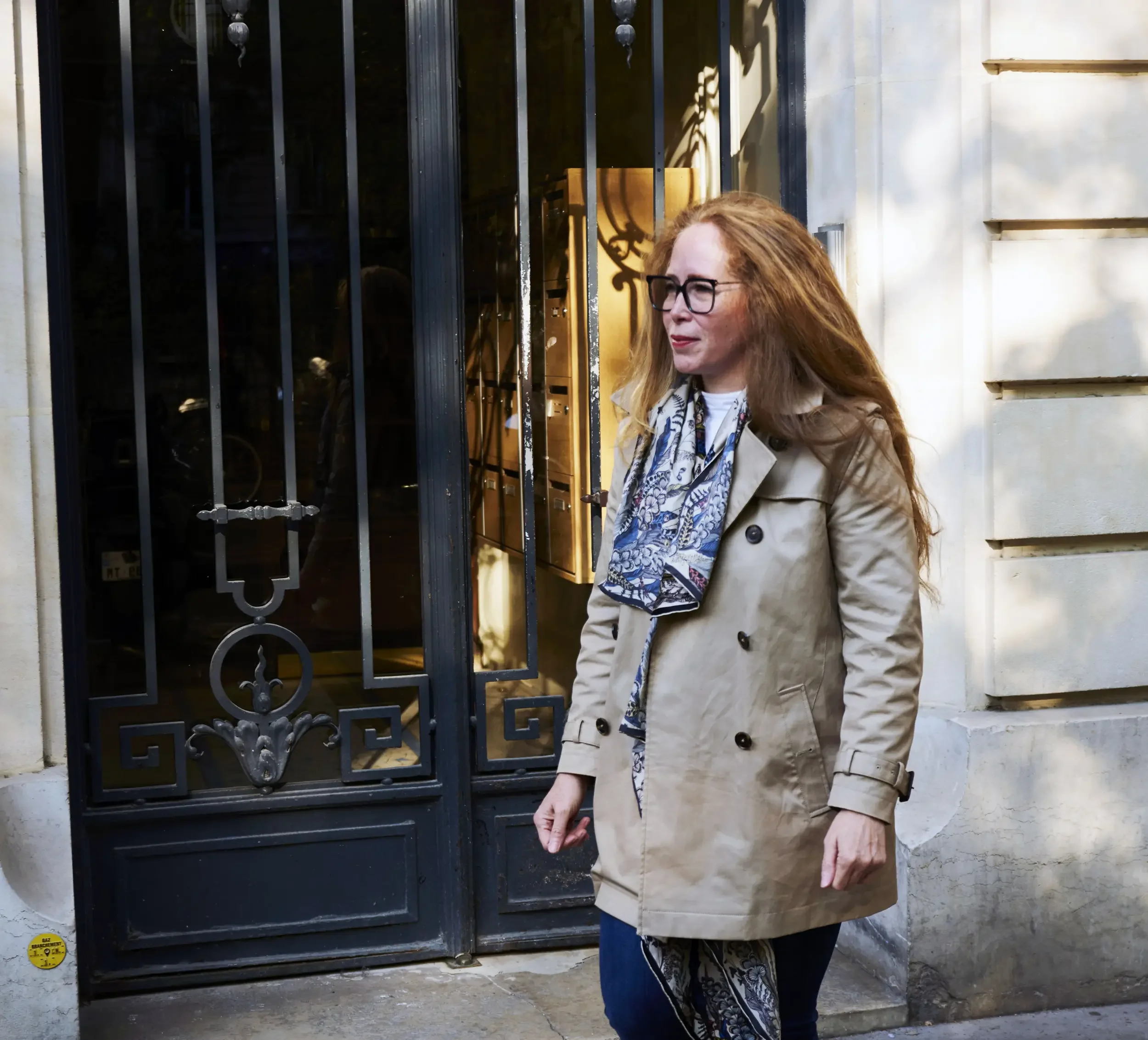 A woman with long red hair, glasses, and a beige trench coat walking past a black wrought iron gate and a building with light-colored stone walls.