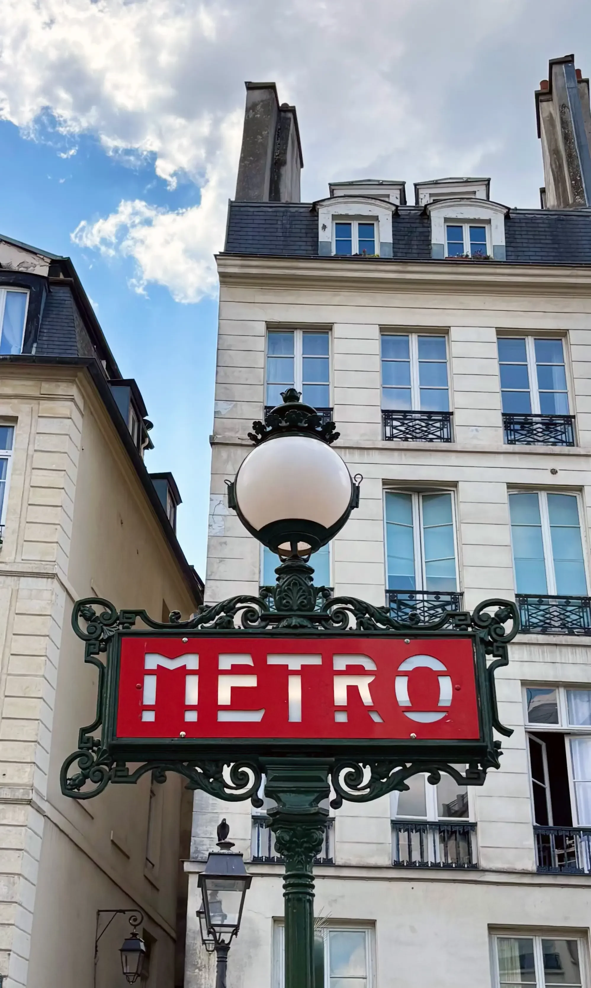 Paris Metro sign with ornate green frame, red background, and white letters, set against historic buildings and a partly cloudy sky.