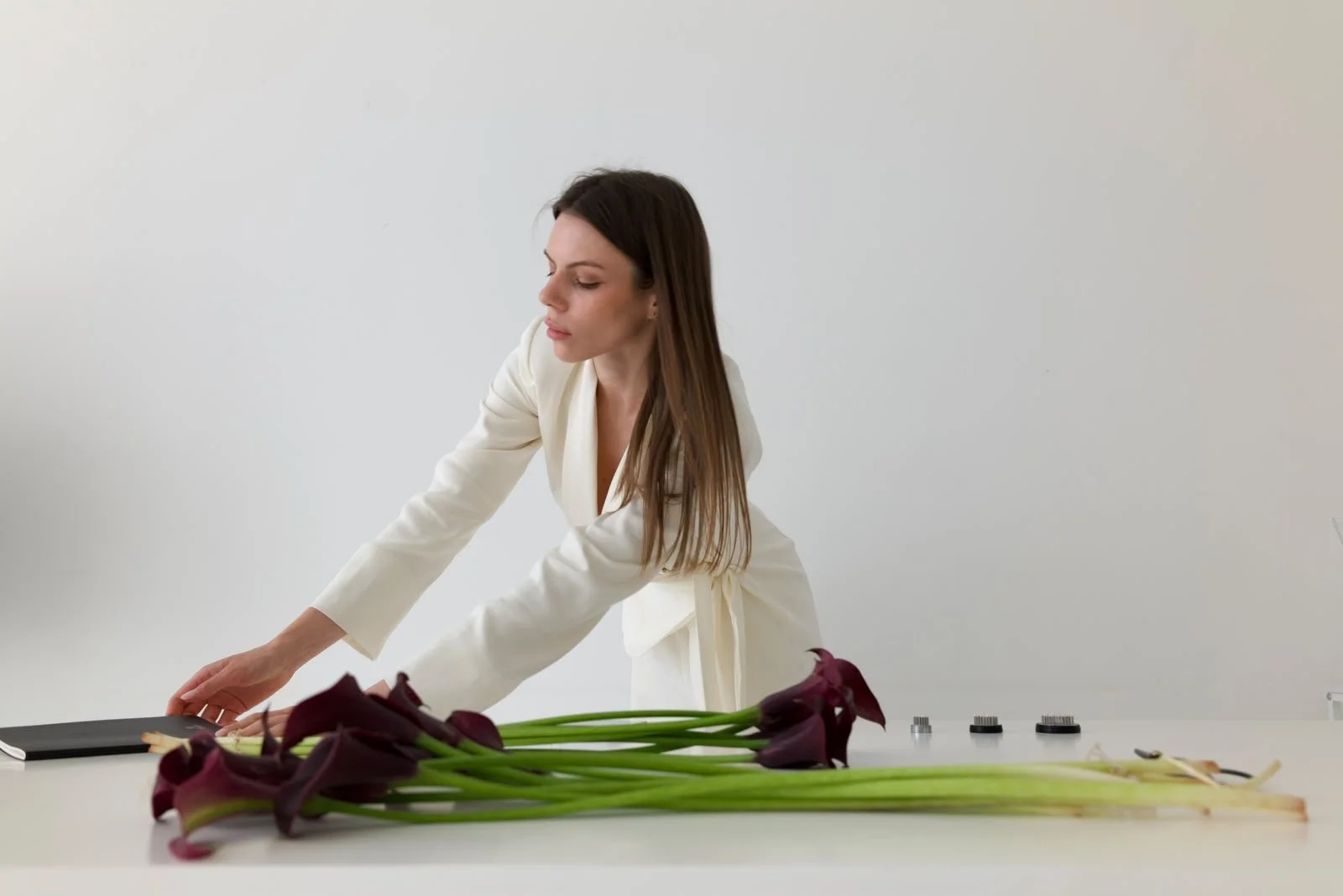 A woman in a white blazer arranging dark purple flowers on a white table.