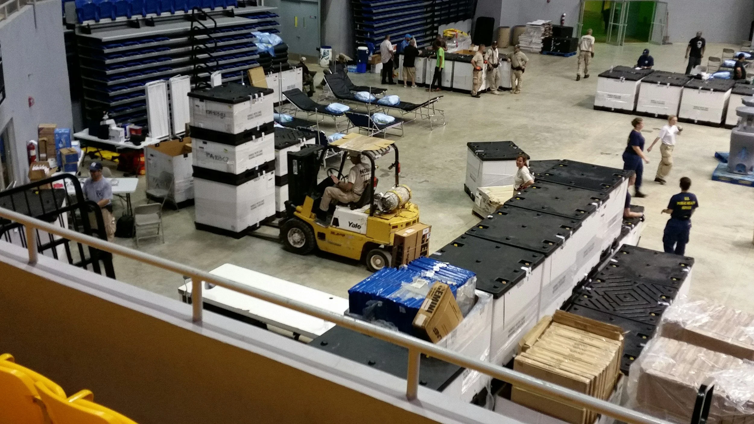 A warehouse with people working, including a person operating a forklift and others handling boxes and supplies.