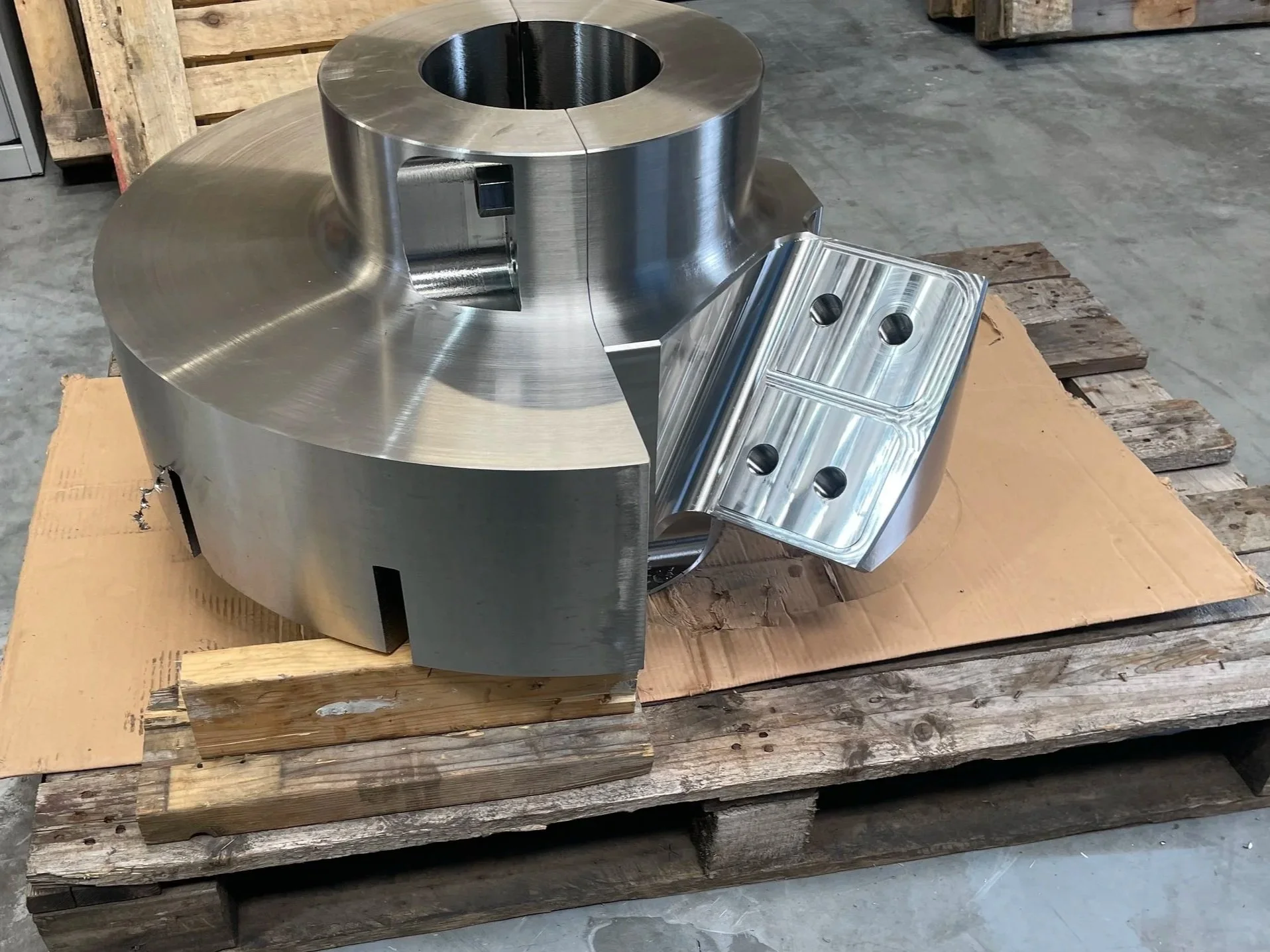 Large metallic mechanical component with cylindrical and rectangular sections, resting on a wooden pallet in an industrial setting.