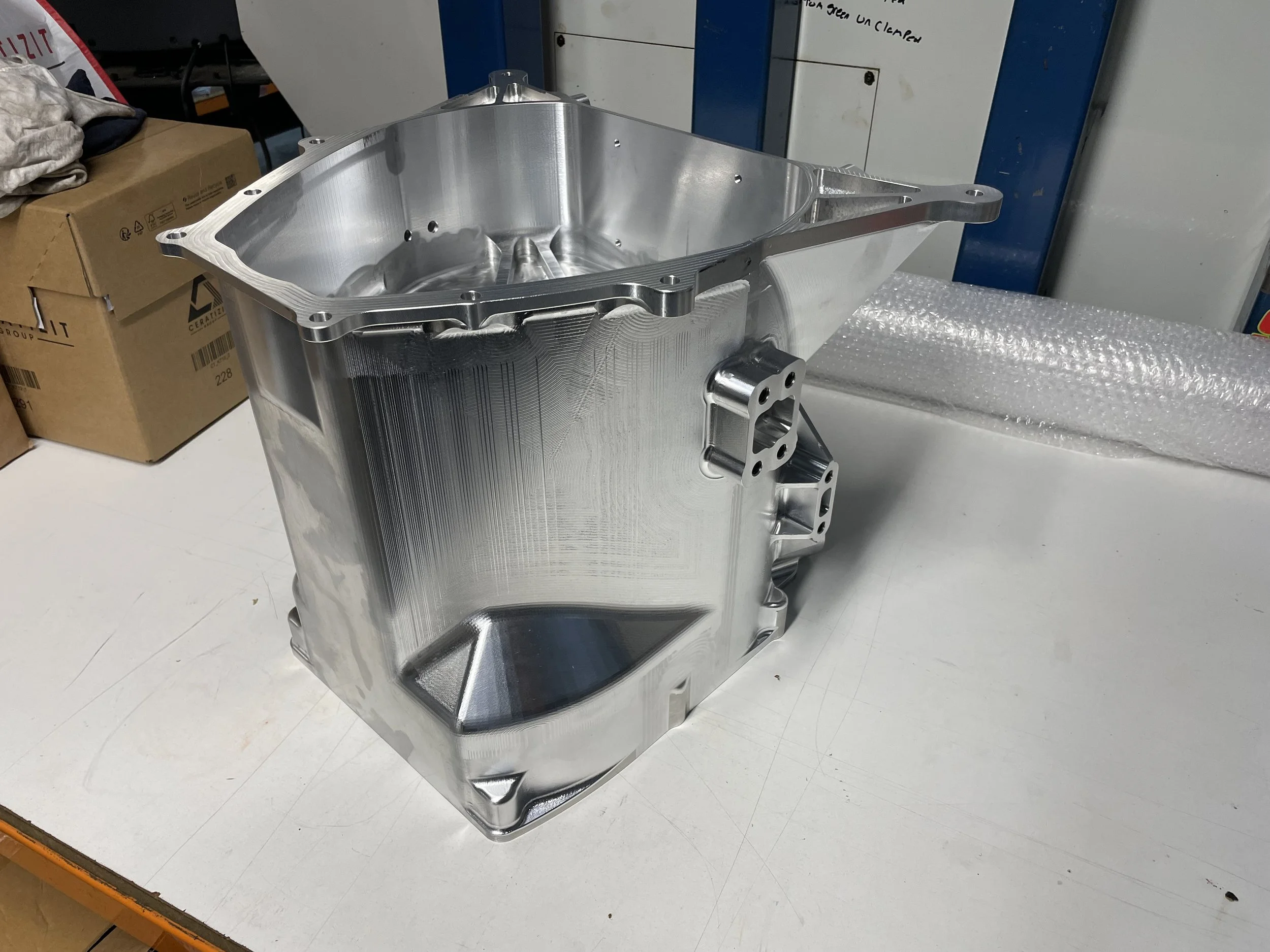 A large, shiny, metallic engine block or machinery component placed on a white work surface in a workshop or manufacturing environment. There are cardboard boxes and bubble wrap nearby.