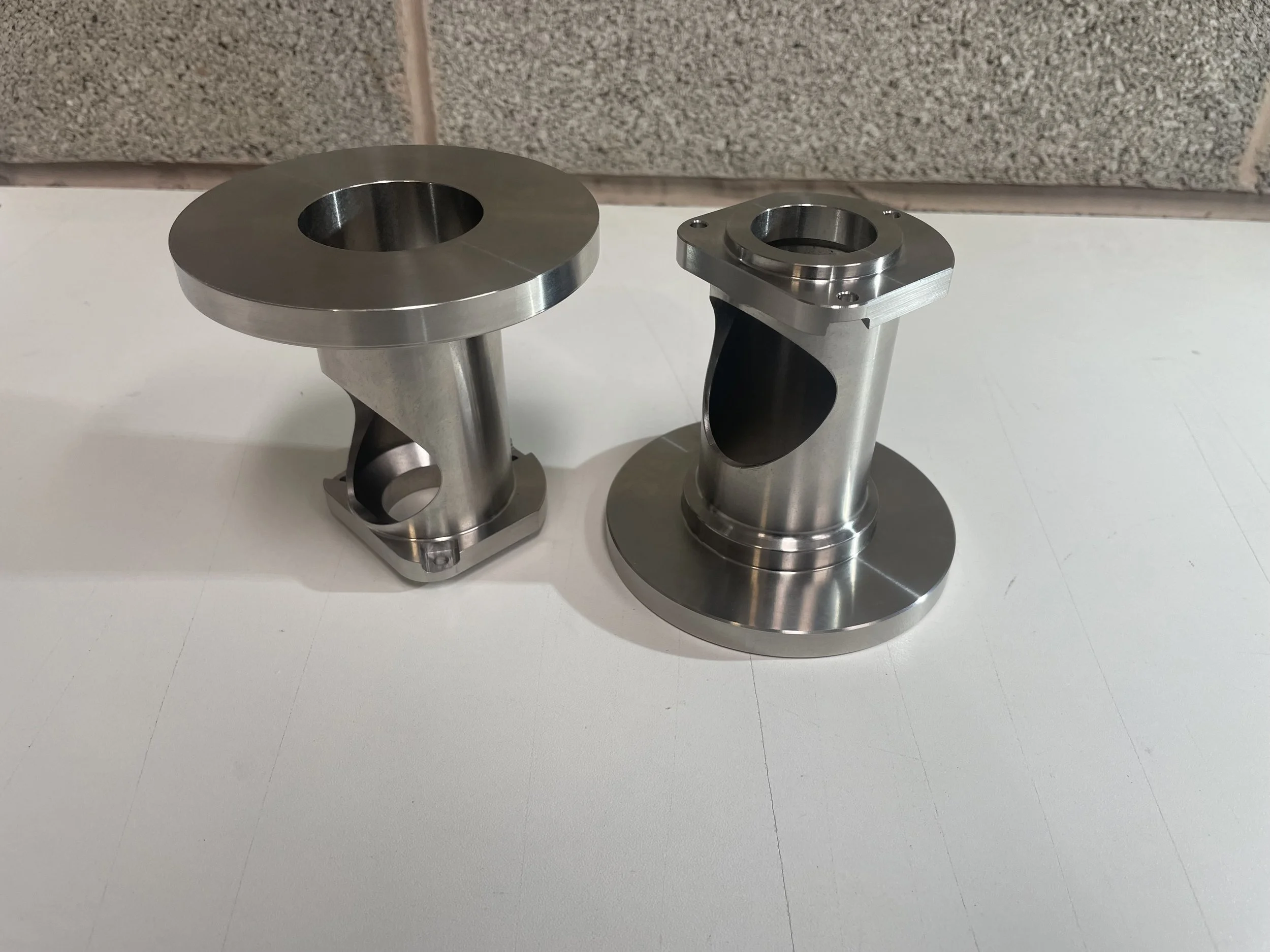 Two polished metal precision machined parts with cylindrical shapes and mounts, on a white surface against a tiled wall background.
