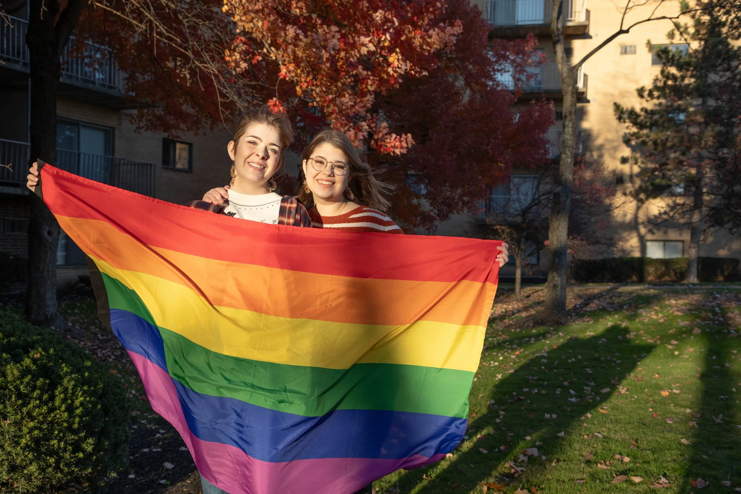 Two young women smiling outdoors holding a rainbow pride flag with arms around one another