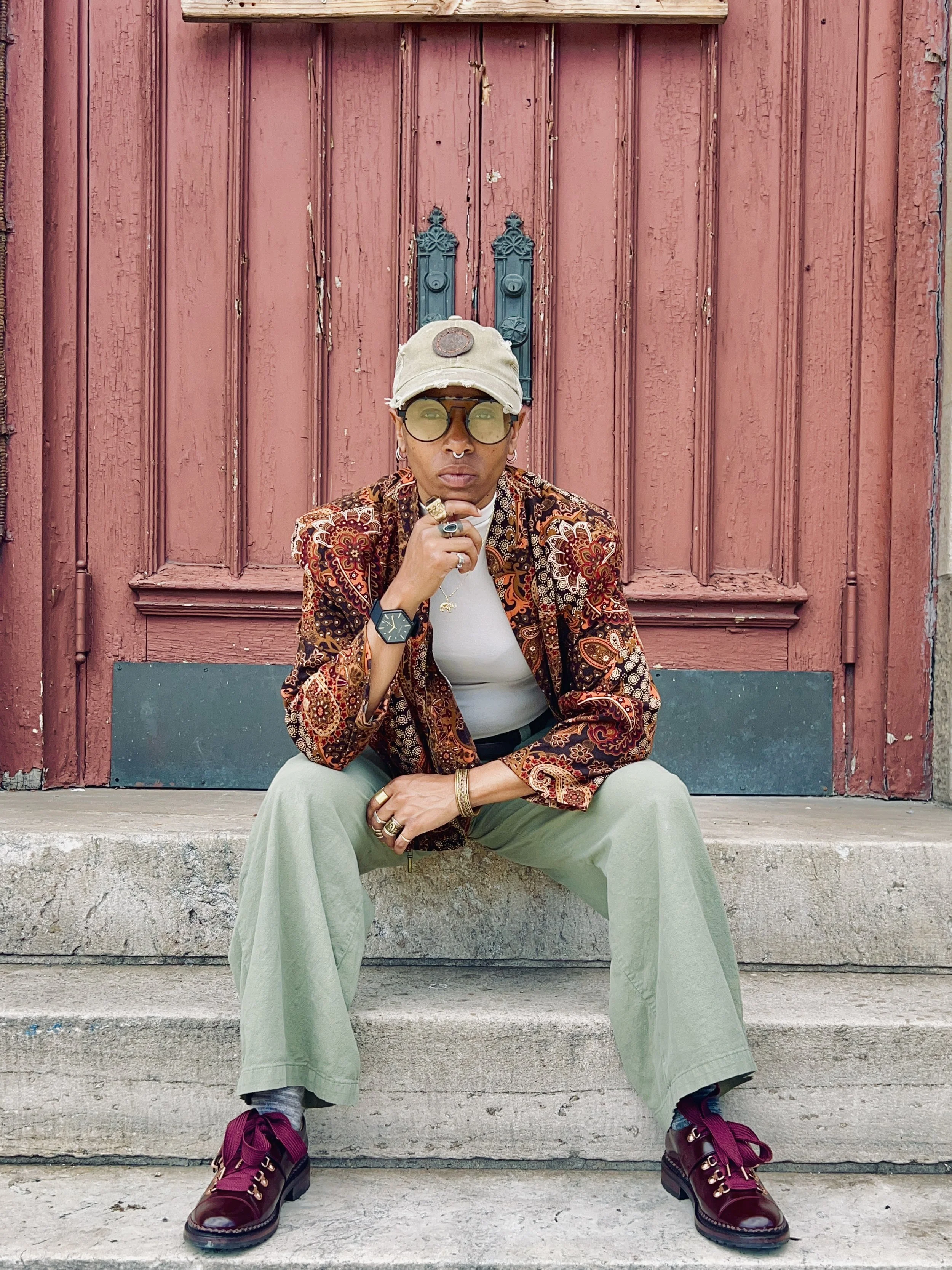 Person sitting on steps in front of a worn red wooden door, wearing a patterned blazer, beige cap, glasses, and burgundy shoes.