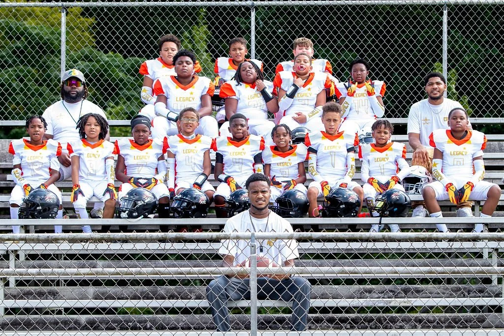 Youth football team and coaches sitting on bleachers at a football field, wearing team uniforms and helmets.