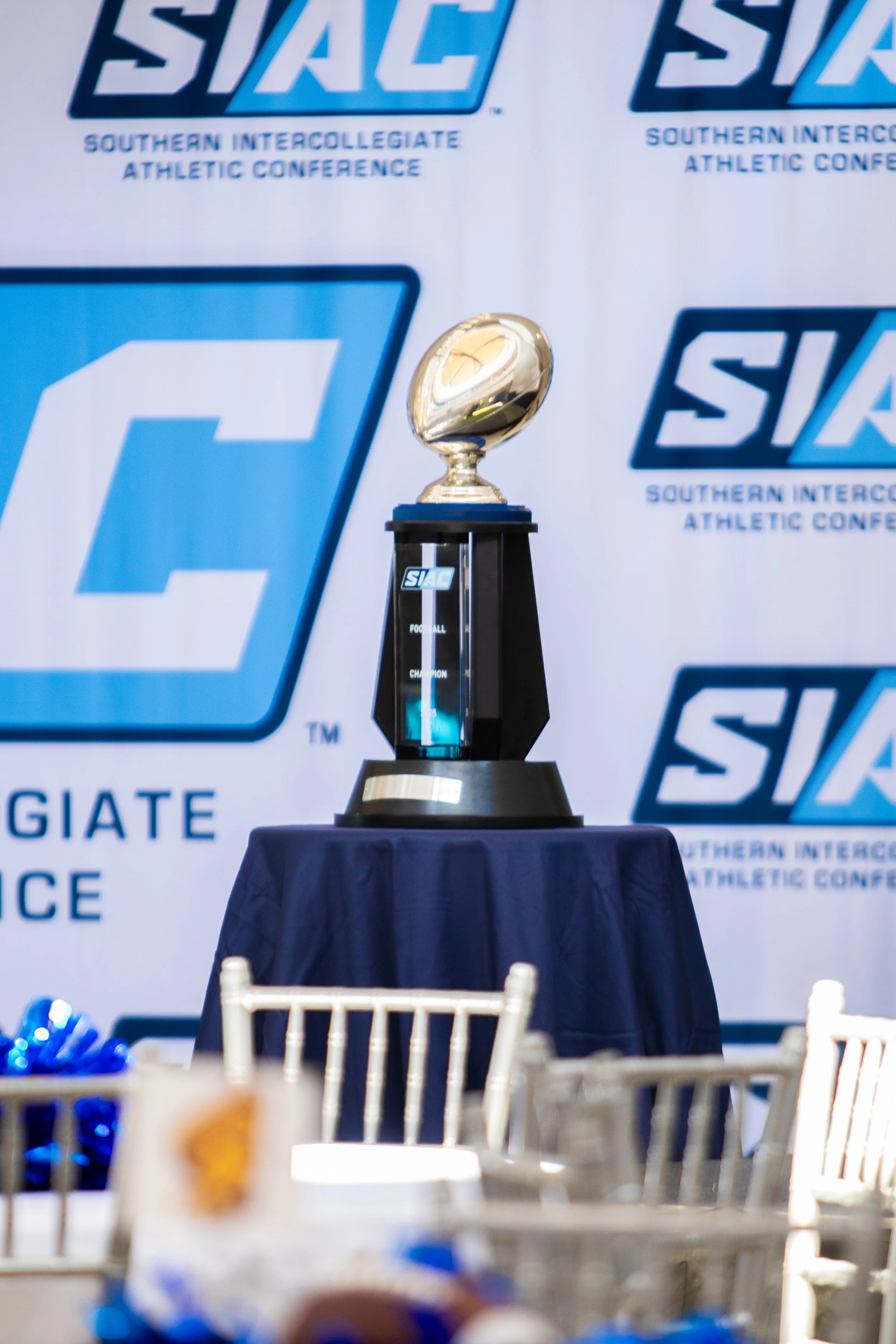 A trophy on a table at an athletic conference, with a backdrop displaying the conference's logo and name.