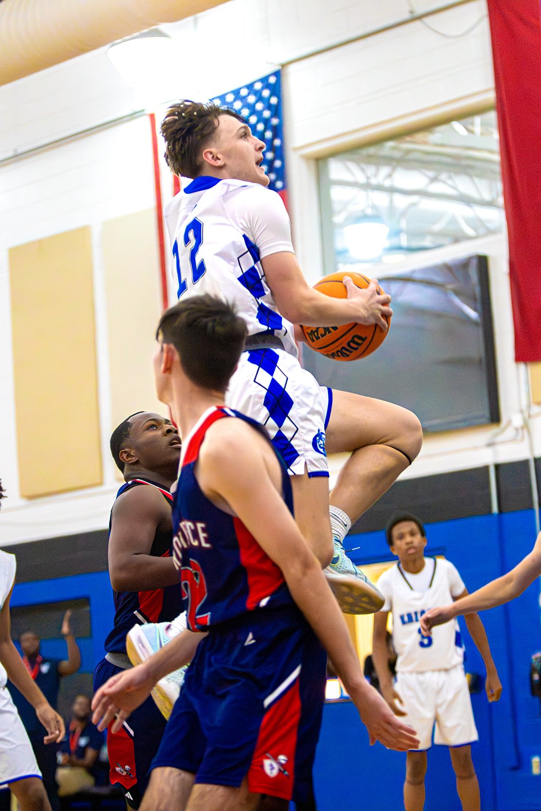 A basketball player in a white and blue uniform jumps up with the ball, attempting a shot during a game. Two players in dark blue and red uniforms are trying to block him, while others are watching from below.