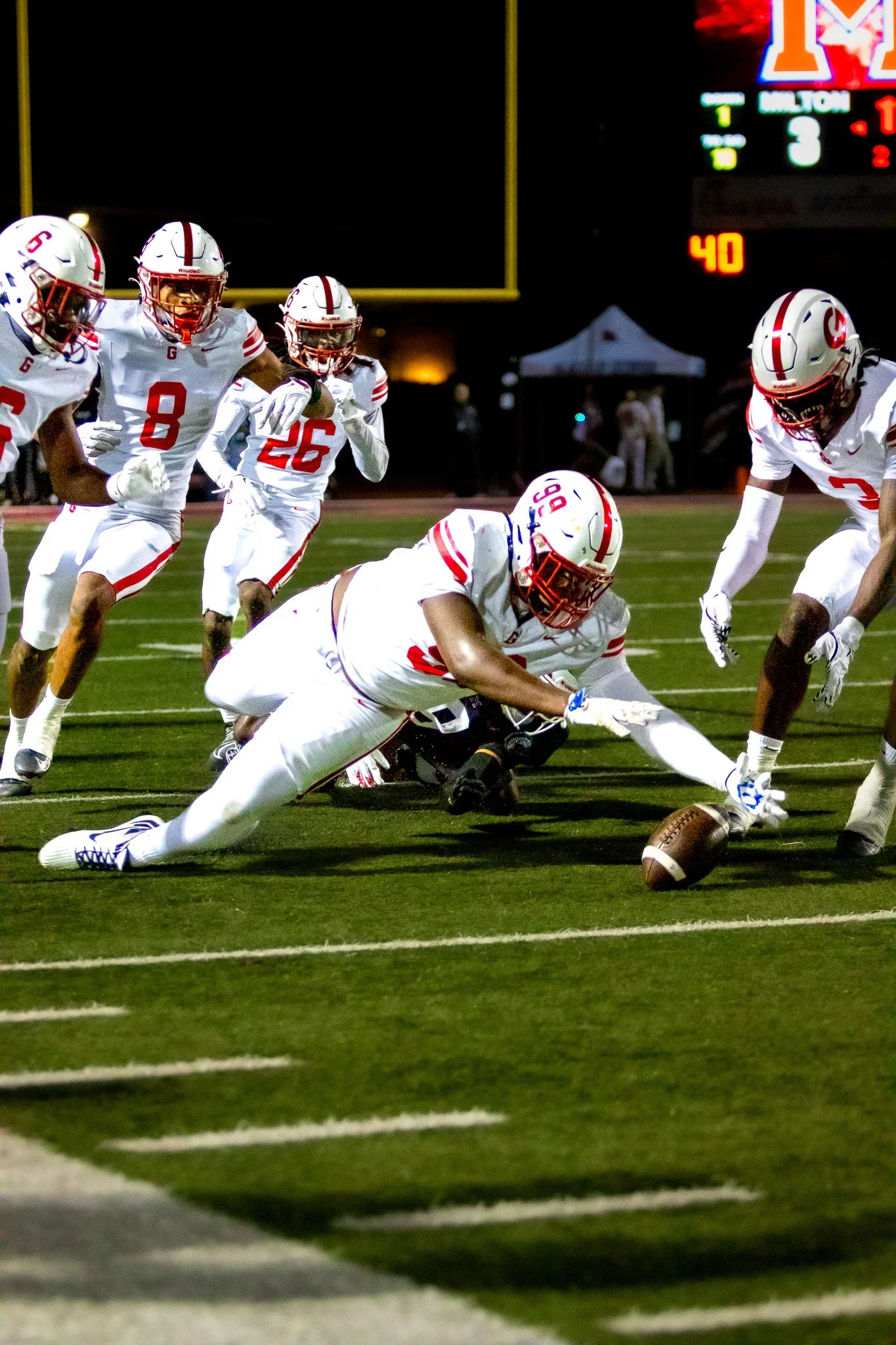 A football player in a white uniform reaches out to recover a fumble on the field during a night game.