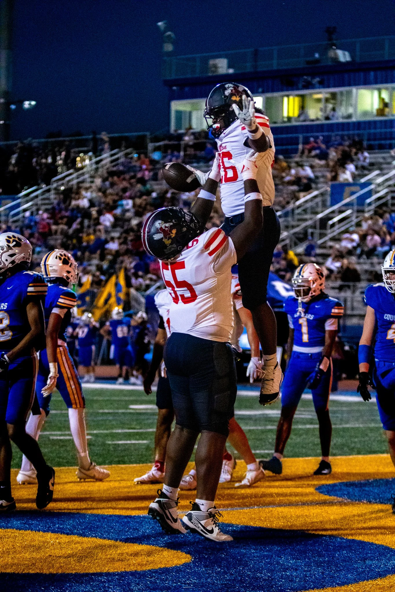 Two football players in football uniforms celebrating on a field, with one player lifted and holding a football, during a game at dusk.