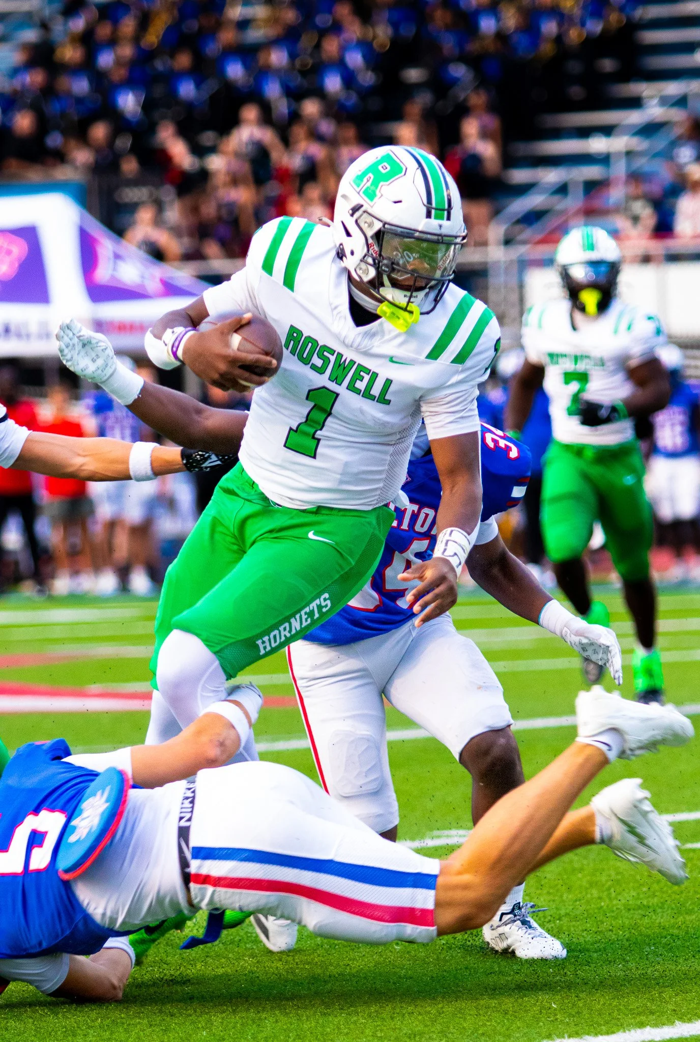 A football player from Roswell High School, wearing a white and green uniform with number 1, is running with the ball while being tackled by a player from another team who is wearing a blue uniform with white pants. Spectators are visible in the background.