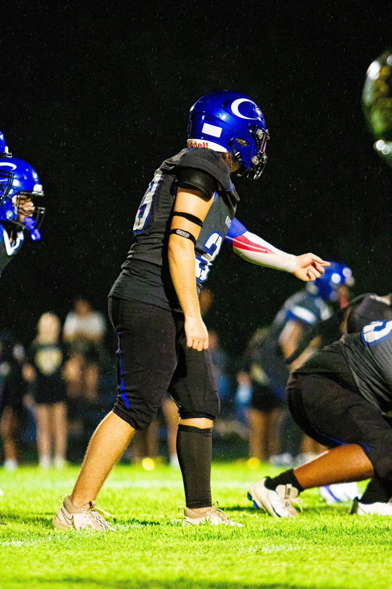 A football player standing on the field during a night game, wearing a black uniform with blue and white accents, a blue helmet with a white logo, and white cleats, with other players and spectators in the background.