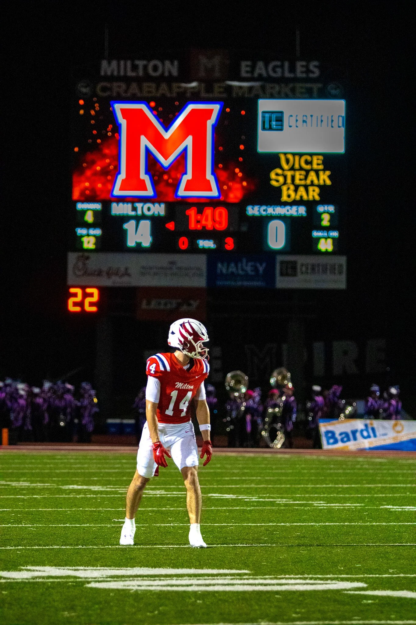A football player wearing a red jersey with the number 14, standing on a football field at night, with a large scoreboard in the background.