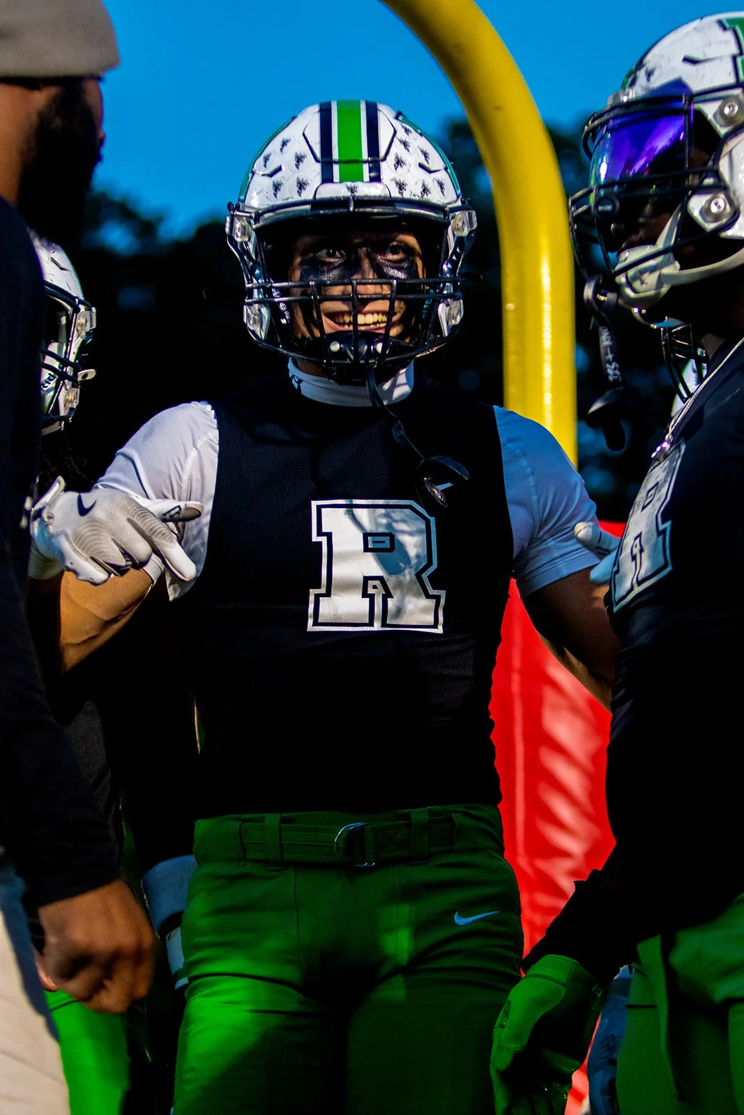 A football player wearing a black jersey with a large white letter R, a white shirt underneath, green pants, a helmet with a green stripe, and black face paint, smiling and standing between teammates during a game or practice.