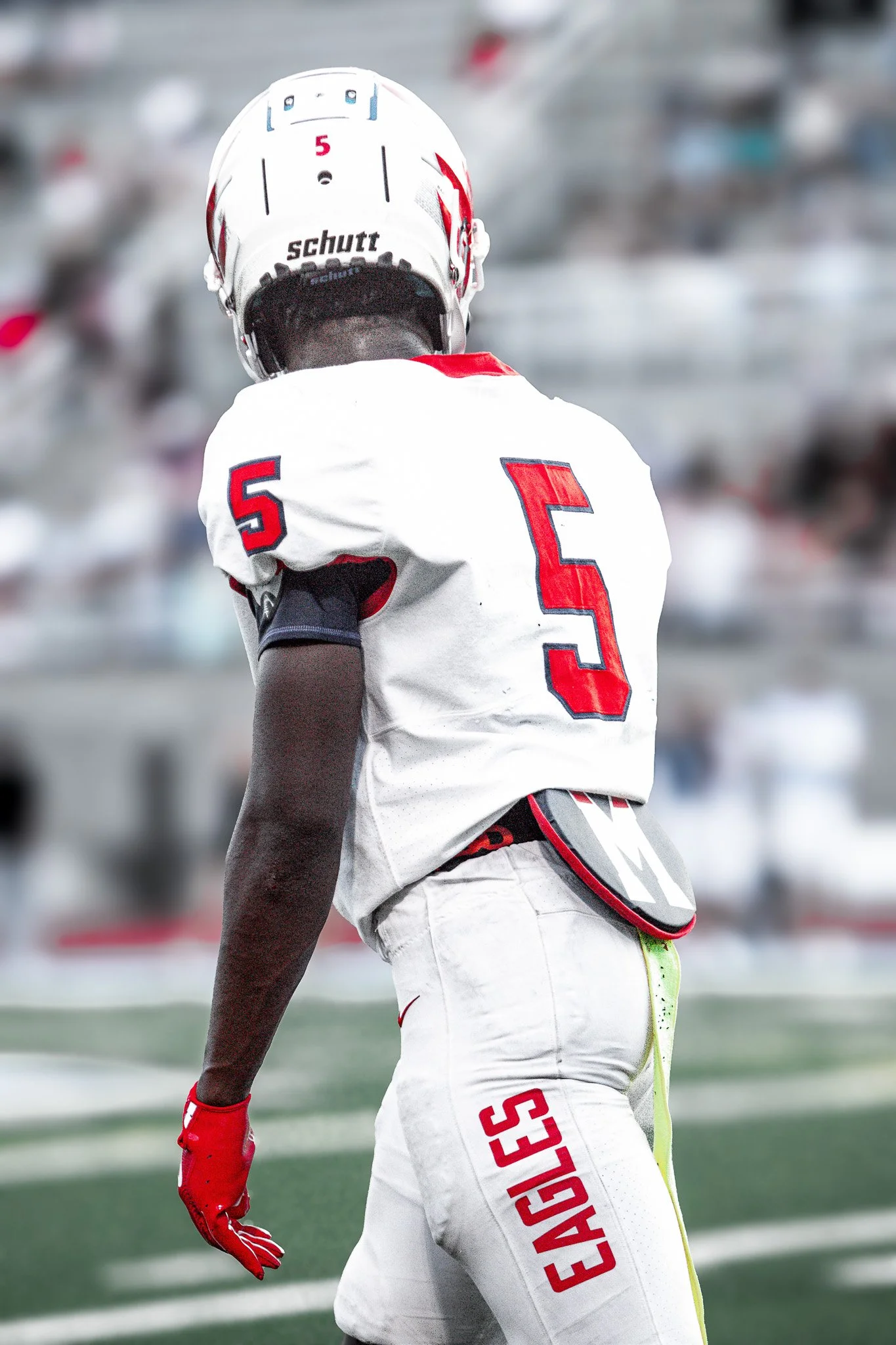 A football player in a white uniform with red and blue details, number 5, standing on a football field with blurred stands in the background.