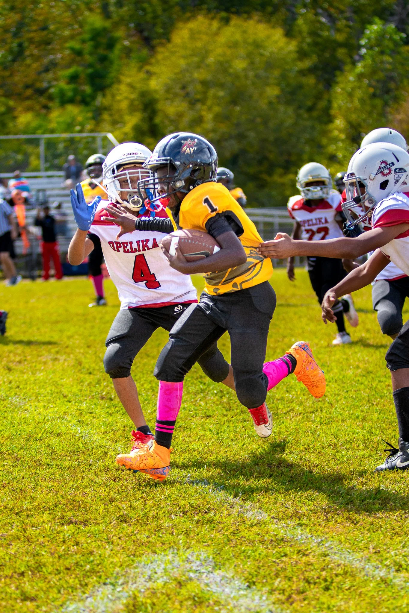 Young football players in action during a game, with one player in a yellow jersey running while holding the football and trying to evade a player in a white jersey attempting to block him on a grassy field.