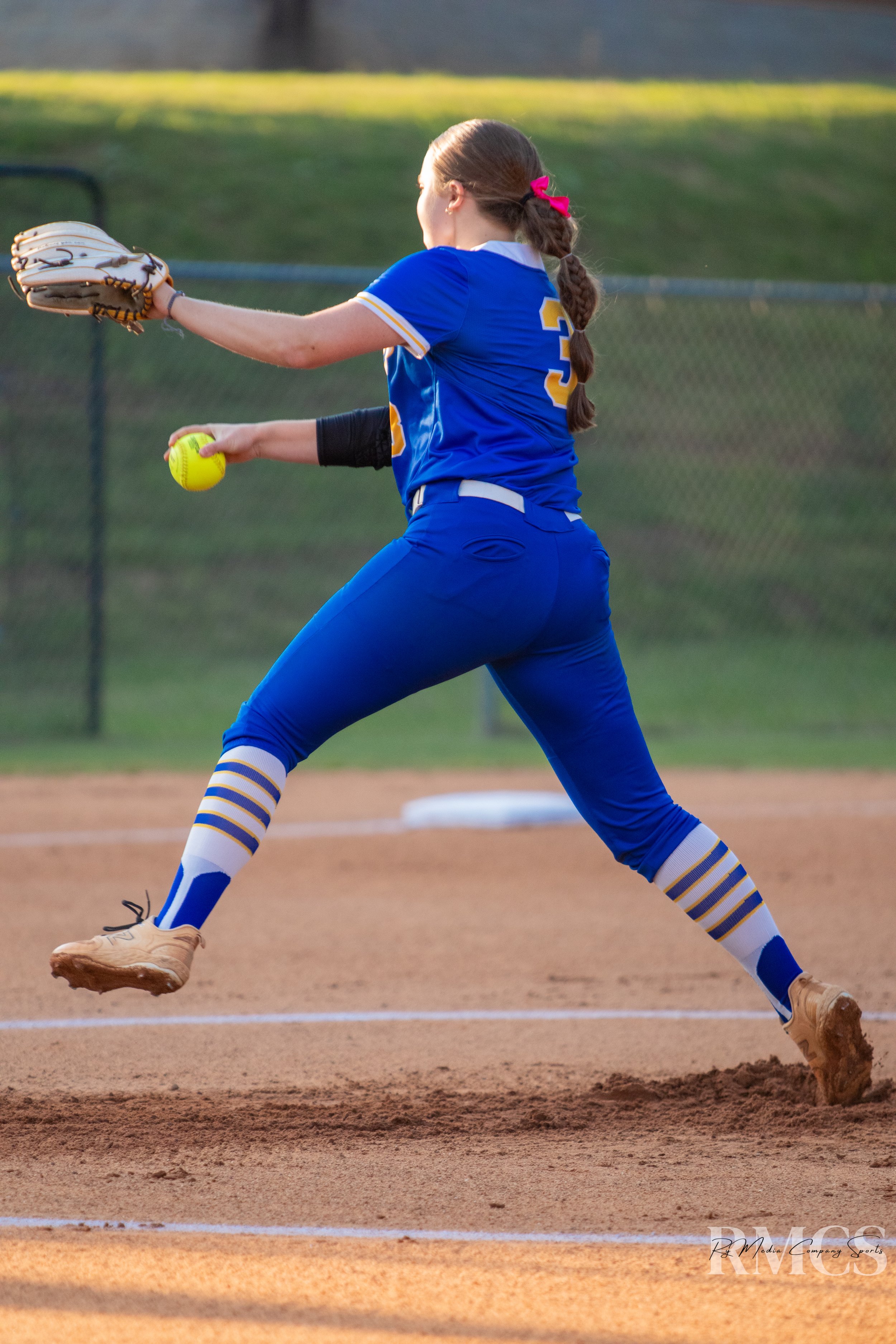 A female softball player wearing a blue uniform with yellow and white accents, fielding a ground ball on the pitcher's mound.