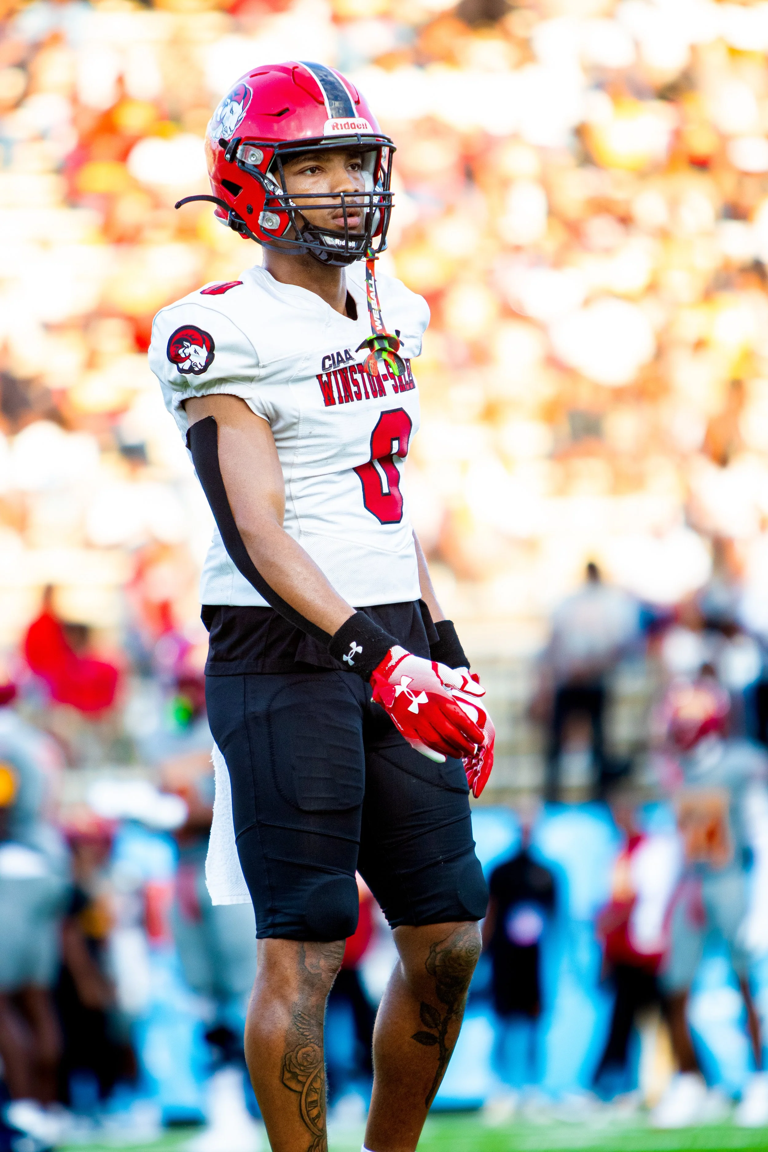 A football player standing on the field in uniform with a red helmet, white jersey with red and black accents, and black shorts, with a crowd in the background.