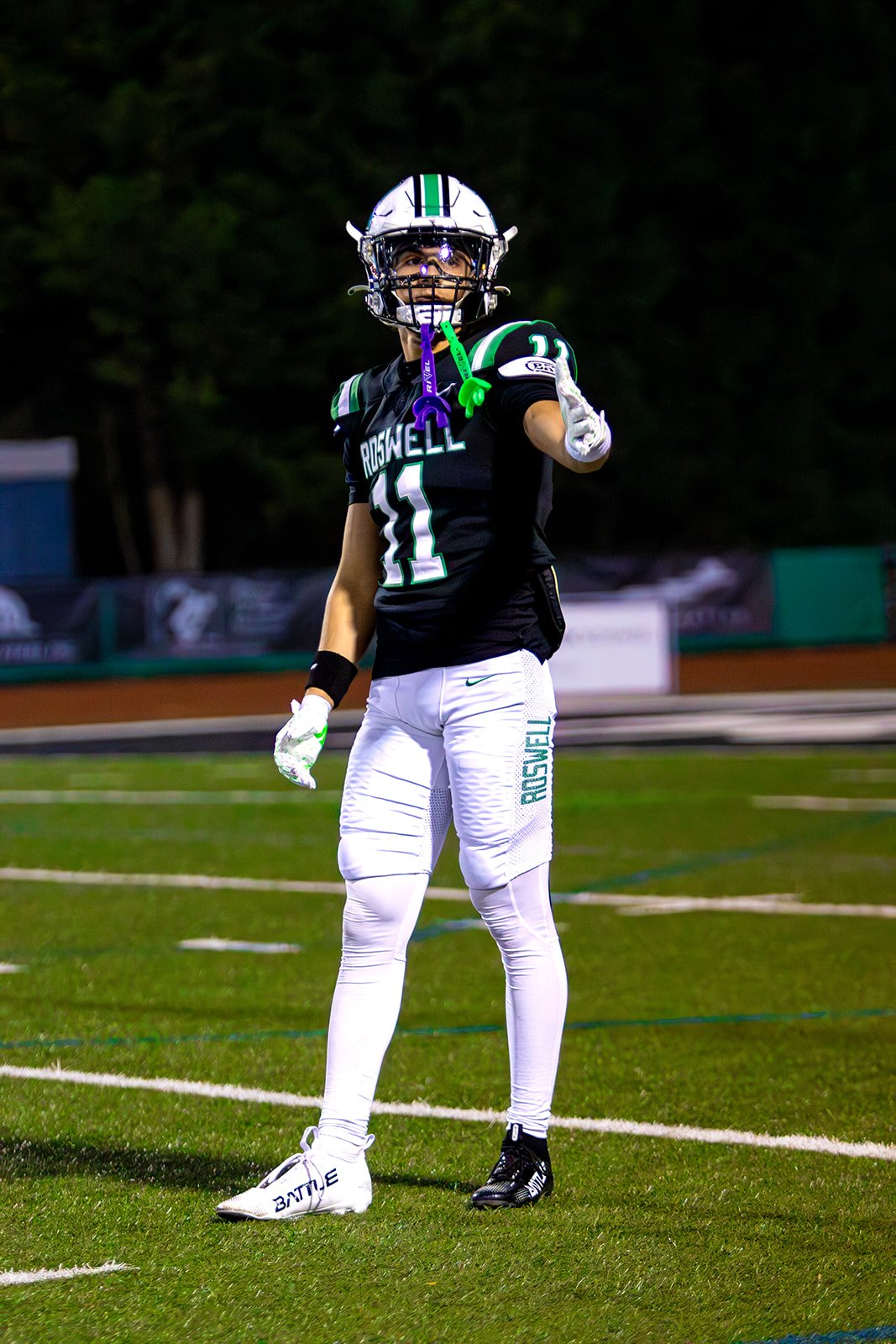 A football player in a black and white uniform with the number 11, standing on a football field, pointing with his right hand, wearing a helmet, gloves, and cleats.
