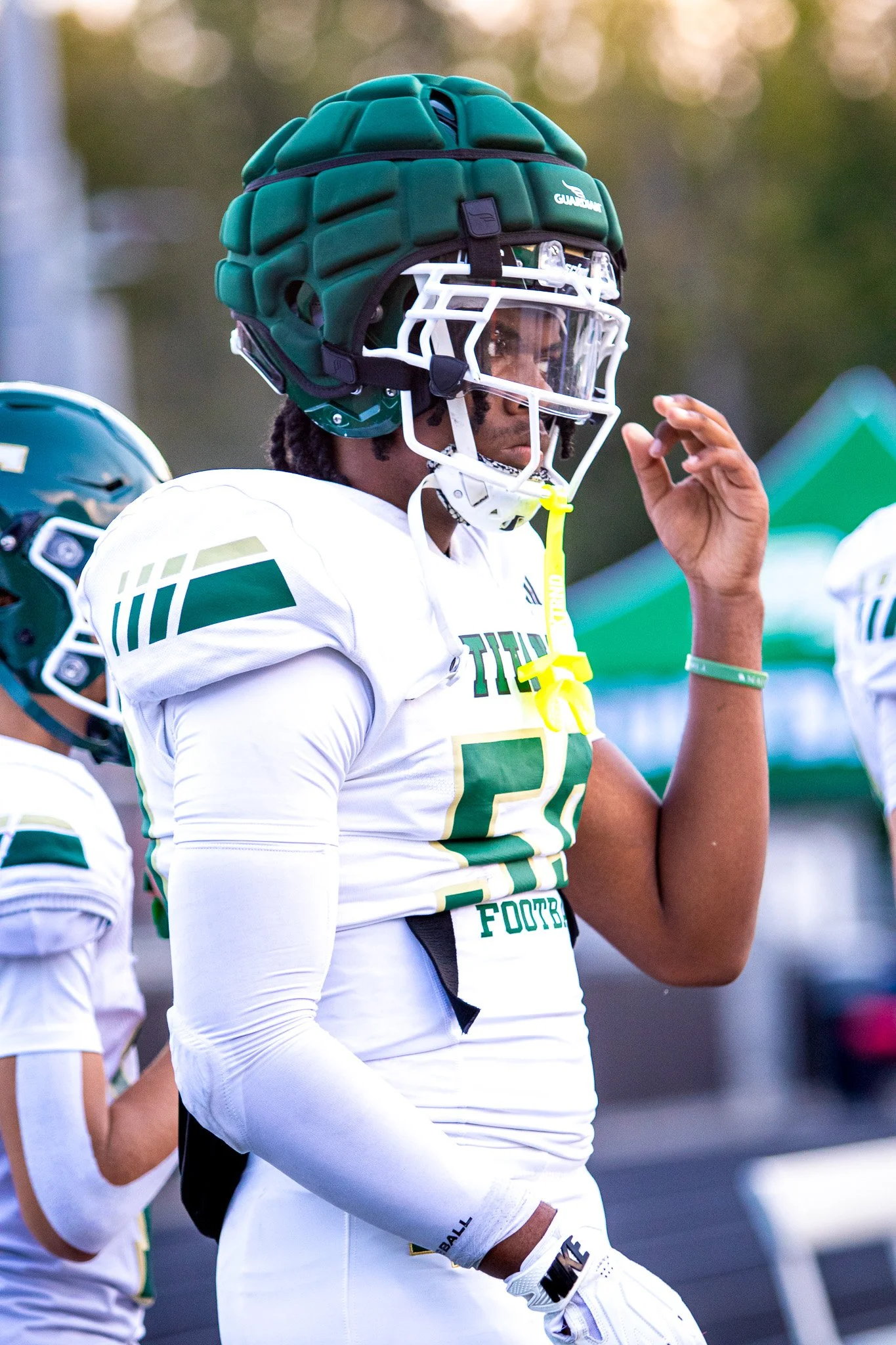 American football player wearing a green helmet, white jersey with green accents, and protective gear, standing with hand raised.