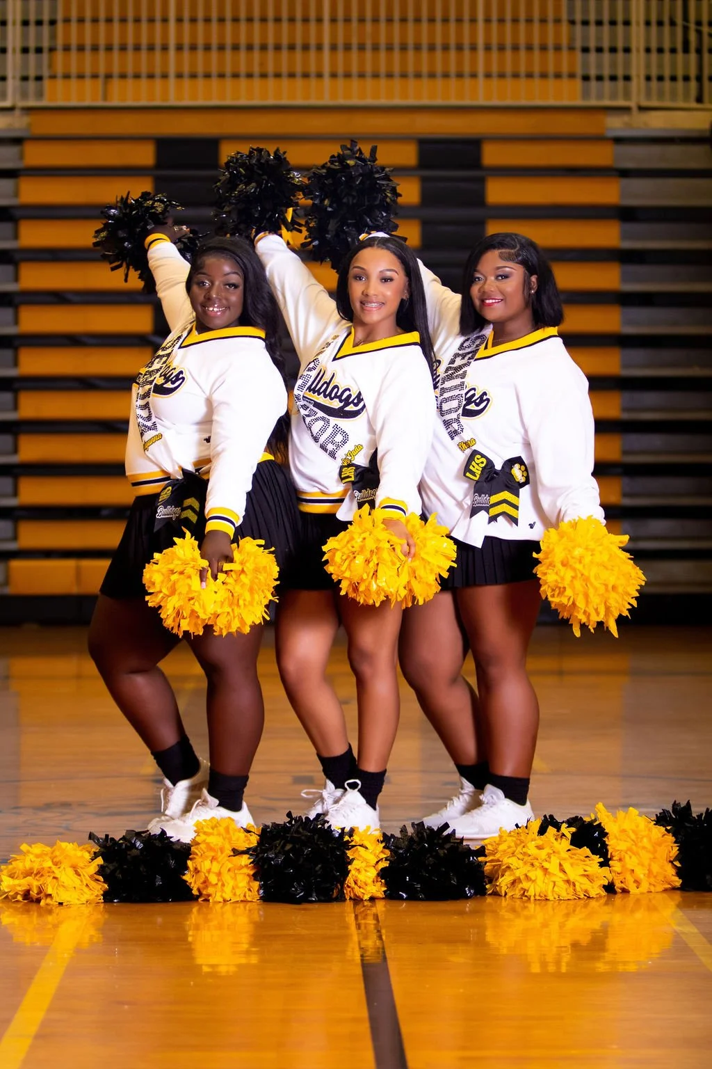 Three cheerleaders in yellow, black, and white uniforms posing with pom-poms in a gymnasium.