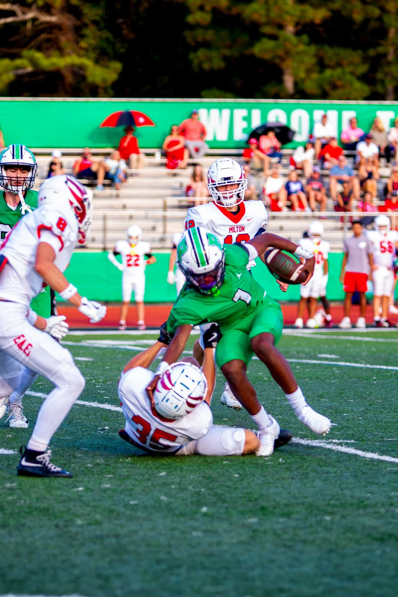 Football players in action during a game, with one player in a green uniform running with the ball while being tackled by an opponent in white, on a football field with spectators in the background.