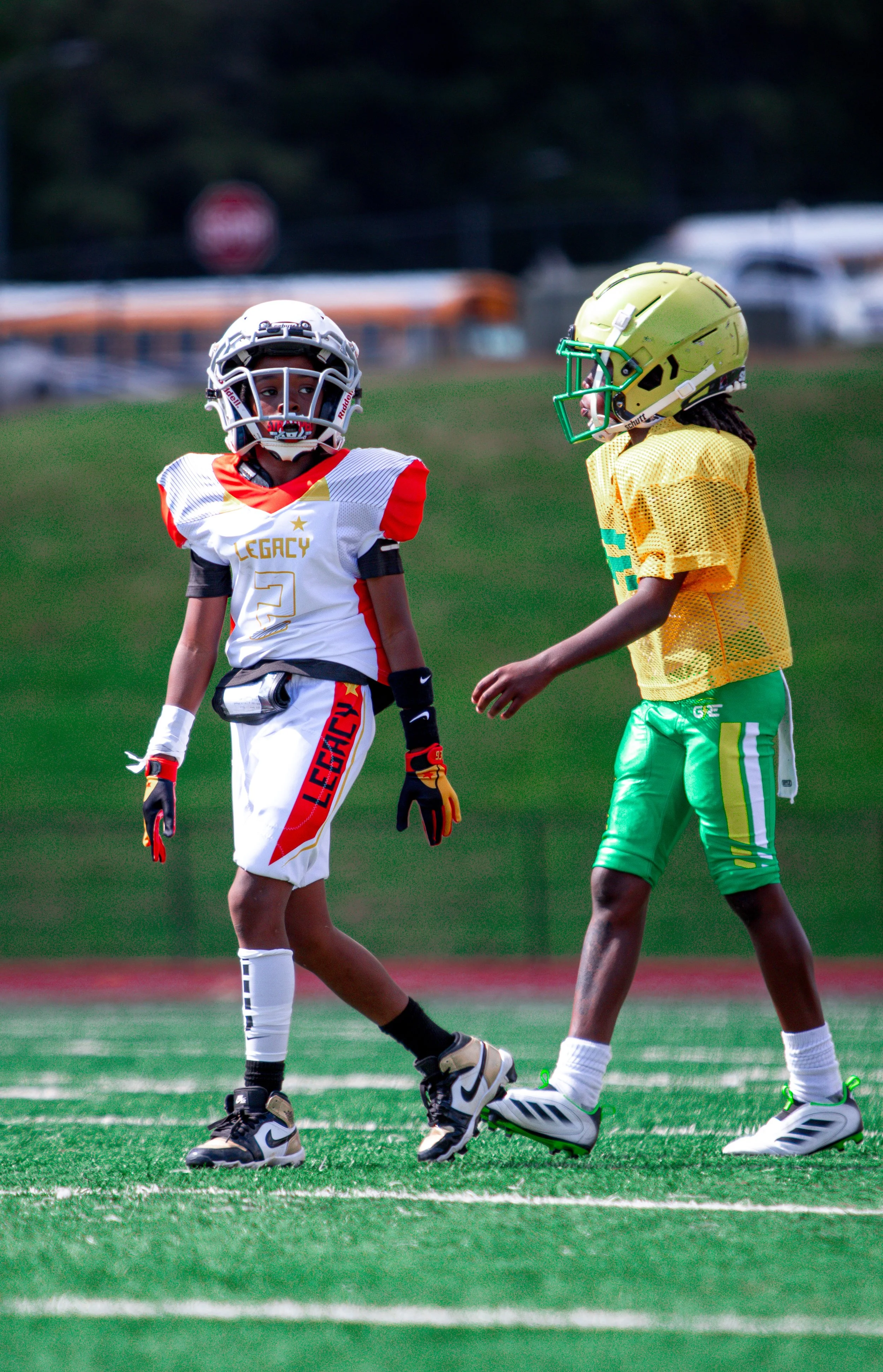 Two young football players wearing helmets and football gear, standing on a green football field during daylight.