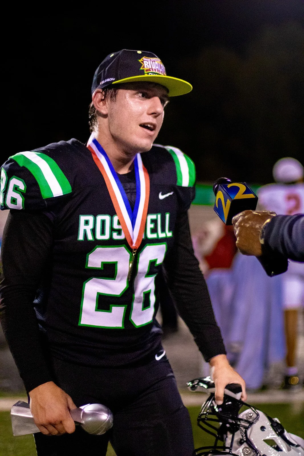 A young football player wearing a black jersey with green and white accents, holding a trophy in one hand, and wearing a medal around his neck, being interviewed by a reporter with a microphone, on a football field at night.