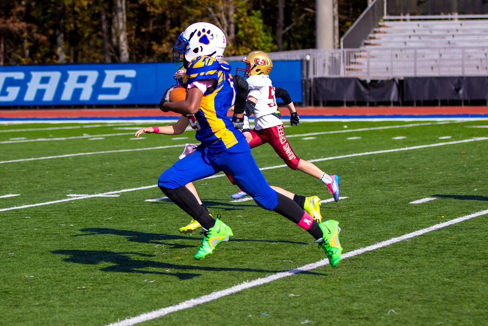 Youth football players running on a field during a game, wearing colorful uniforms and helmets.