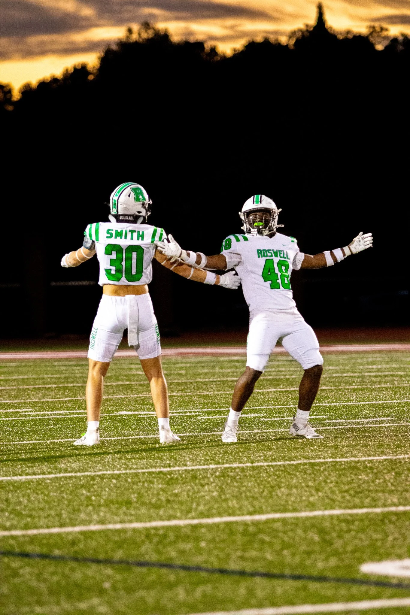 Two football players in white and green uniforms celebrating on the field during sunset.
