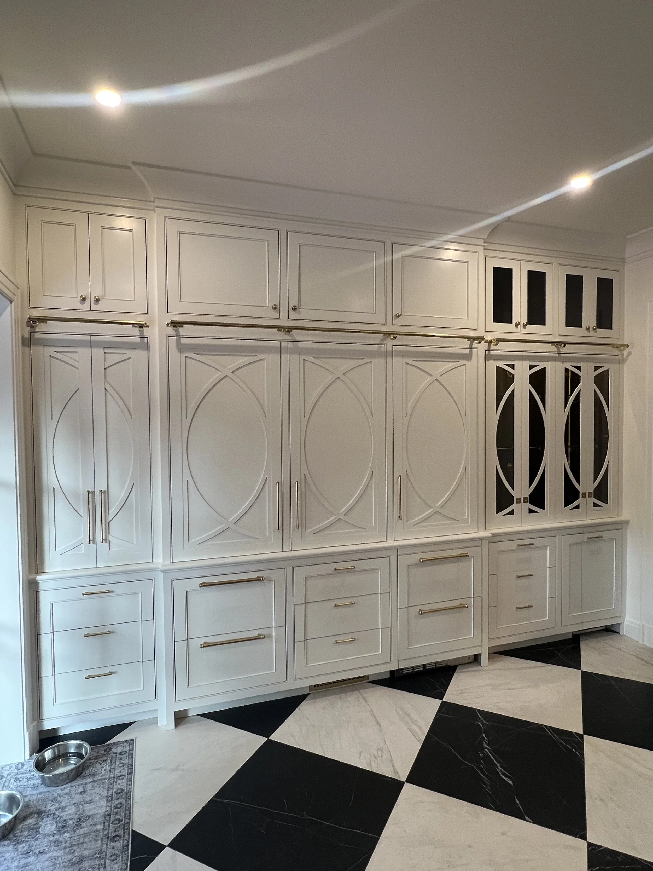 White kitchen cabinets with gold handles and black glass-paneled doors, black and white checkered floor tiles, two stainless steel pet bowls on a small rug, and a pet food mat
